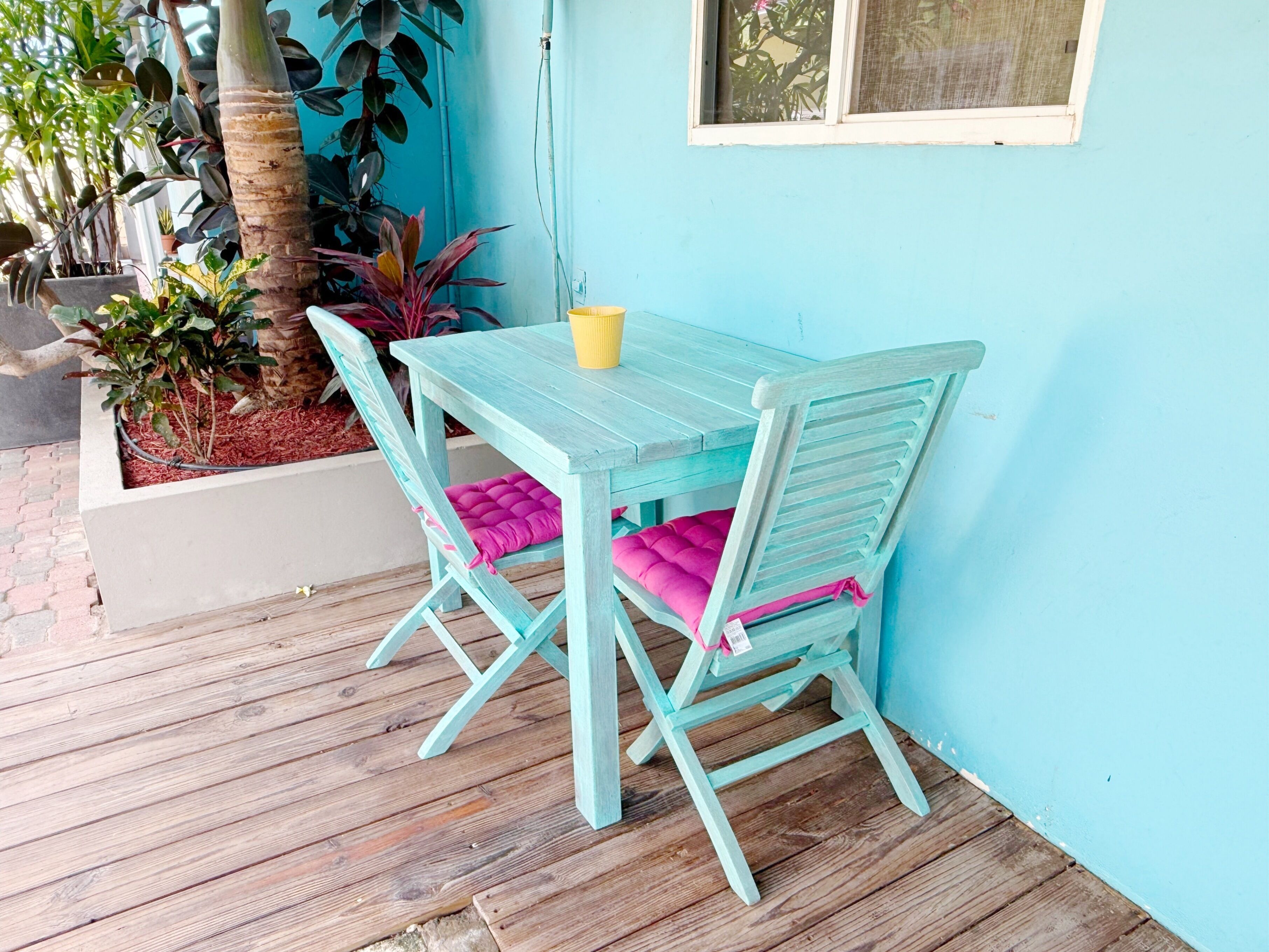 Een lichtblauwe houten tafel met twee stoelen, een gele beker en een plantenpot met planten.