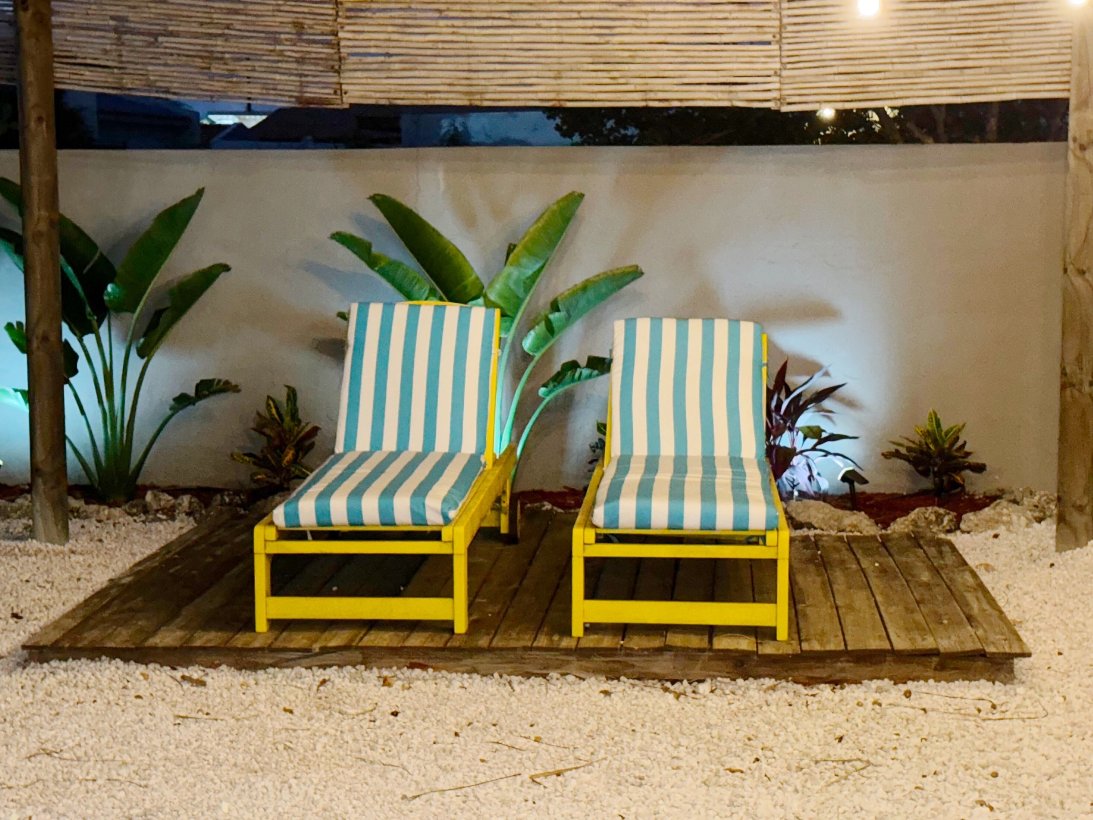 Dos sillones a rayas en una terraza de madera con suelo blanco texturizado, rodeados de plantas tropicales.