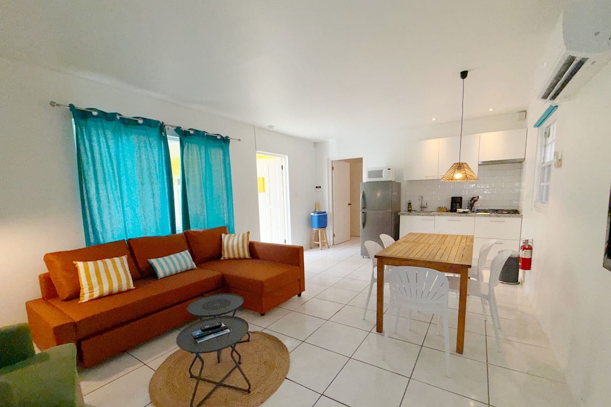 Una sala de estar moderna con sofá, mesa de centro y mesa de comedor. Hay una cocina con refrigerador, microondas y lavamanos.