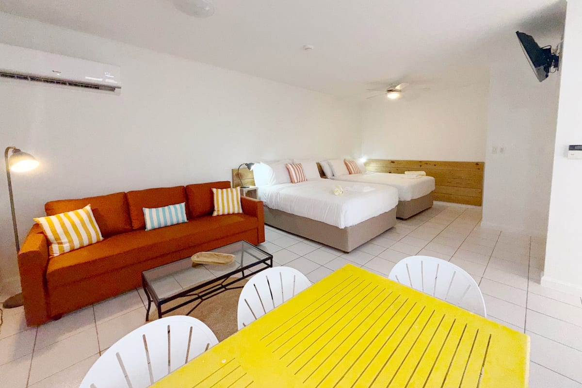 Una habitación de hotel con un sofá, dos camas, una mesa amarilla y sillas blancas.