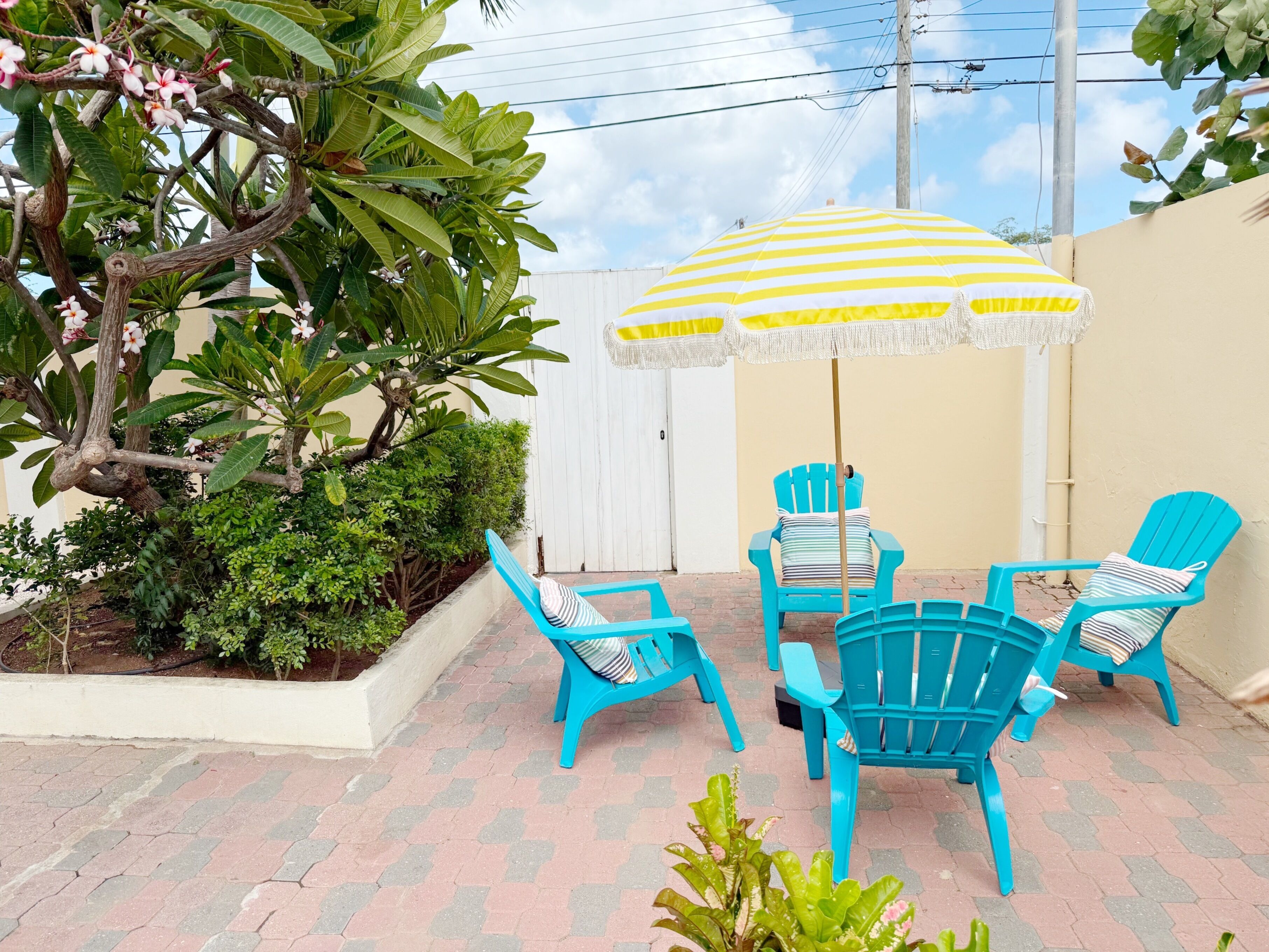 A patio with blue chairs and a yellow-striped umbrella, surrounded by lush greenery and a white wall.