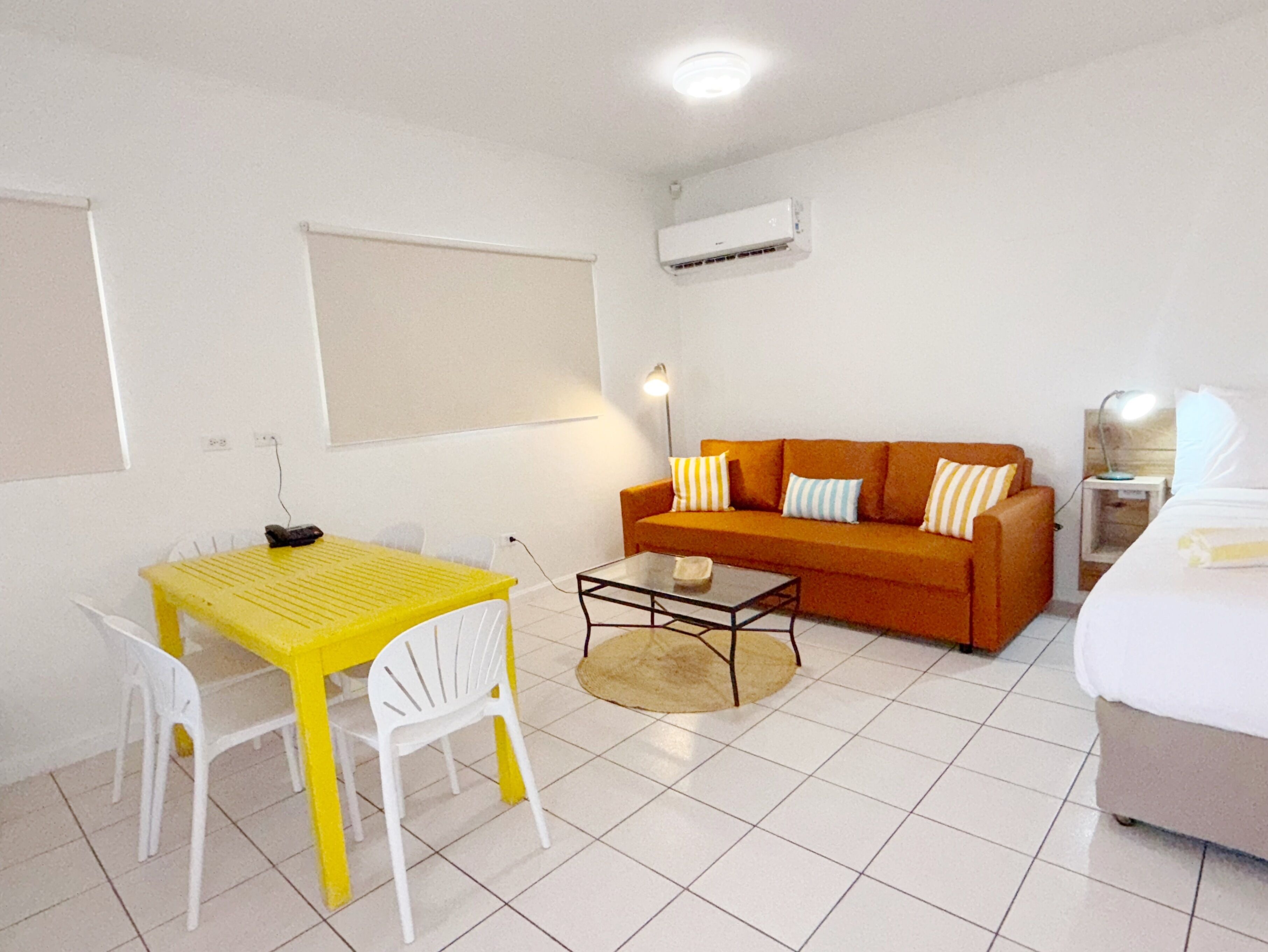 A small, bright room with a yellow table and white chairs, an orange sofa with striped pillows, a small coffee table, a white bed, and an air conditioning unit on the wall.