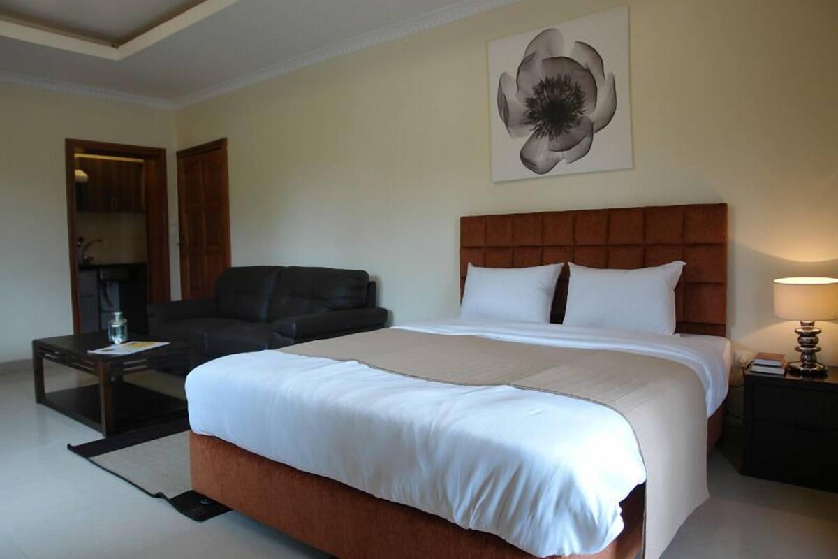 A hotel room with a large bed, a sofa, a coffee table, and a nightstand with a lamp.