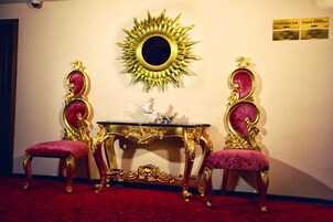 Quarto com duas cadeiras elaboradas, uma mesa pequena e um espelho grande em formato de sol na parede. Há placas indicando um salão de conferências e instalações de ginástica.