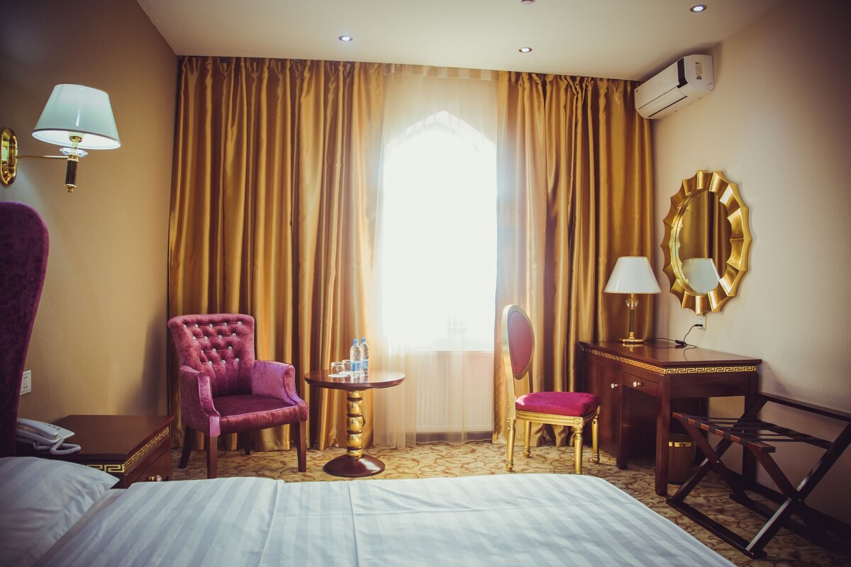 A hotel room with a bed, a purple armchair, a small round table with a lamp, a desk with a chair, and a large mirror.