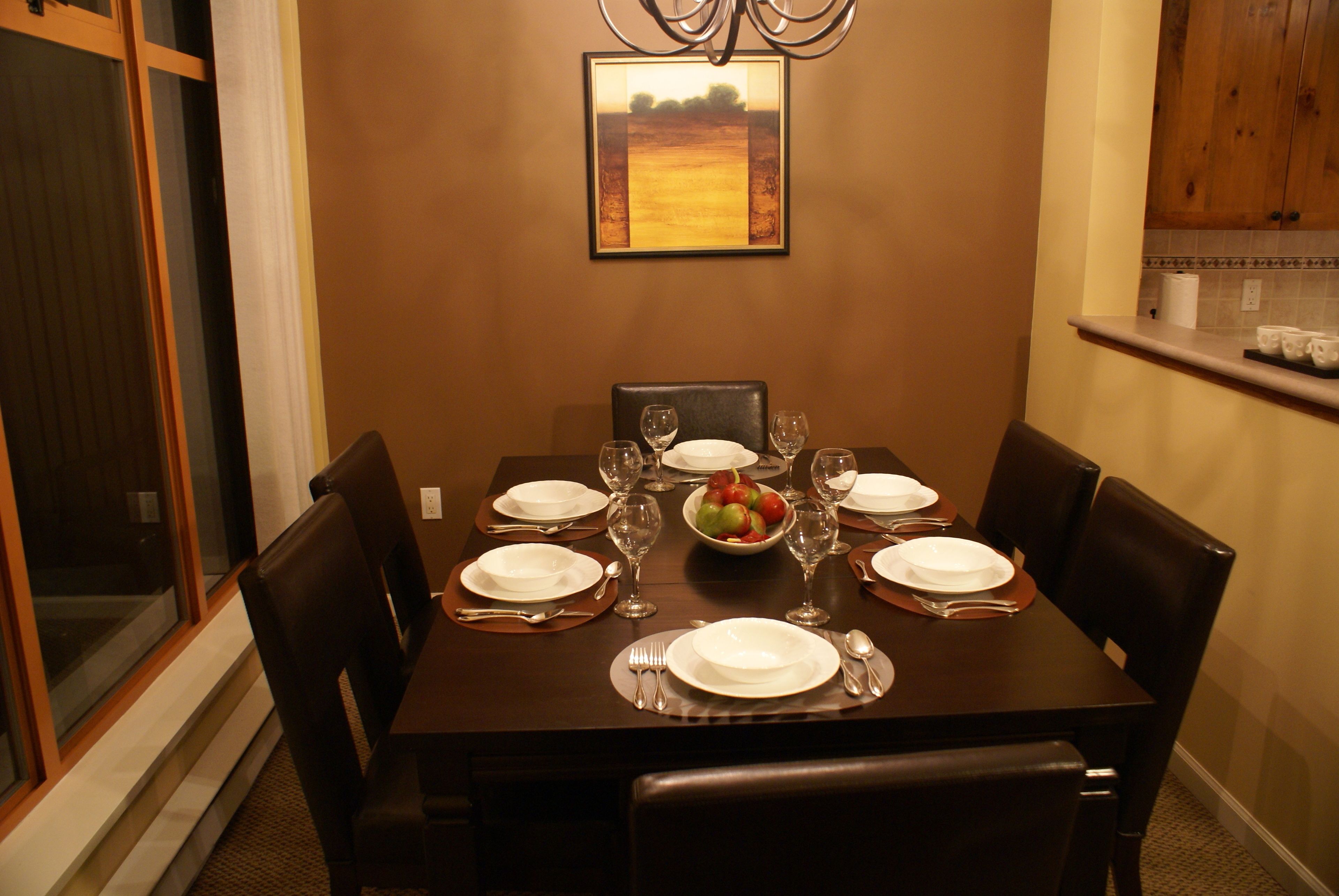 A dining table set for six with plates, cutlery, and wine glasses, a fruit bowl, and a framed picture on the wall.