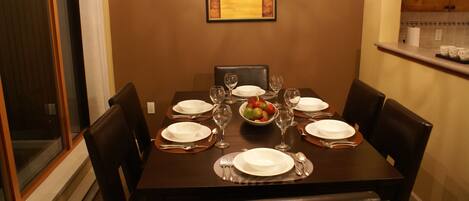 A dining table set for six with plates, cutlery, and wine glasses, a fruit bowl, and a framed picture on the wall.