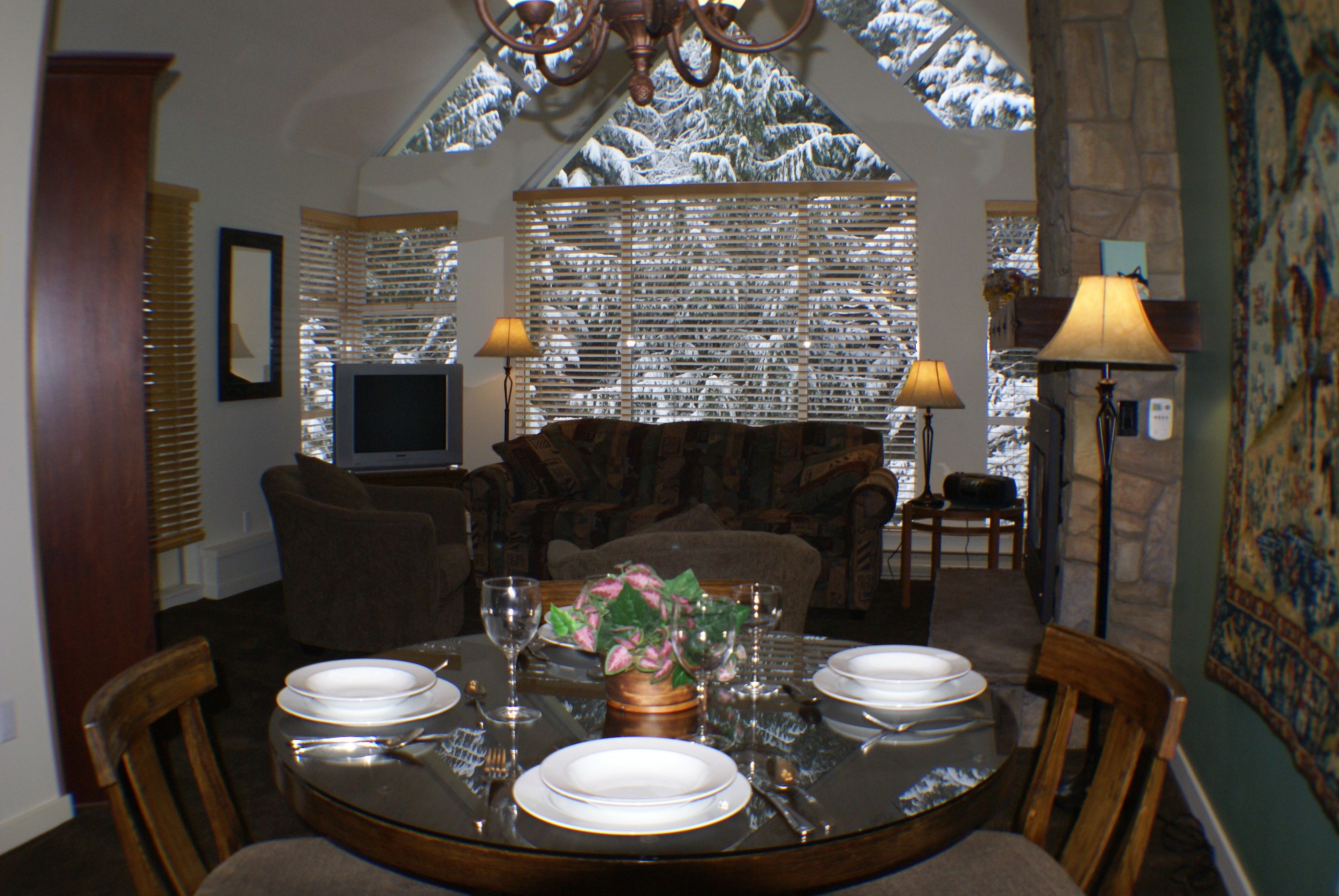 A dining area with a round table set for four, surrounded by chairs. There is a television on a stand to the left and a lamp to the right.