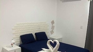A bedroom with a white headboard, a bed with a blue bedspread, a white nightstand with a lamp, and a wall-mounted air conditioner.