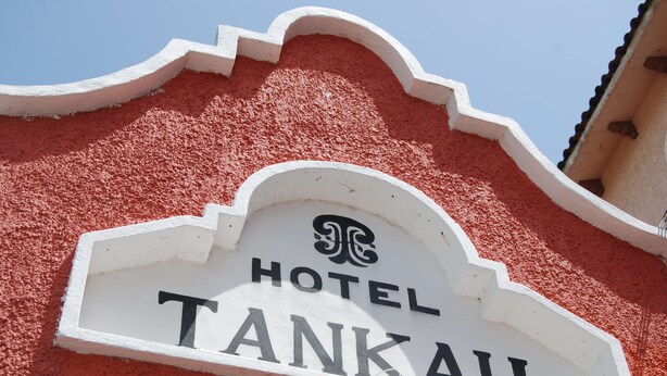 Placa do Hotel Tanka com fundo branco e letras pretas, fixada em uma parede vermelha com textura.