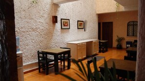Uma área de refeições com mesas e cadeiras de madeira, parede branca com textura e quadros emoldurados, e um espaço de cozinha com armários e pia.