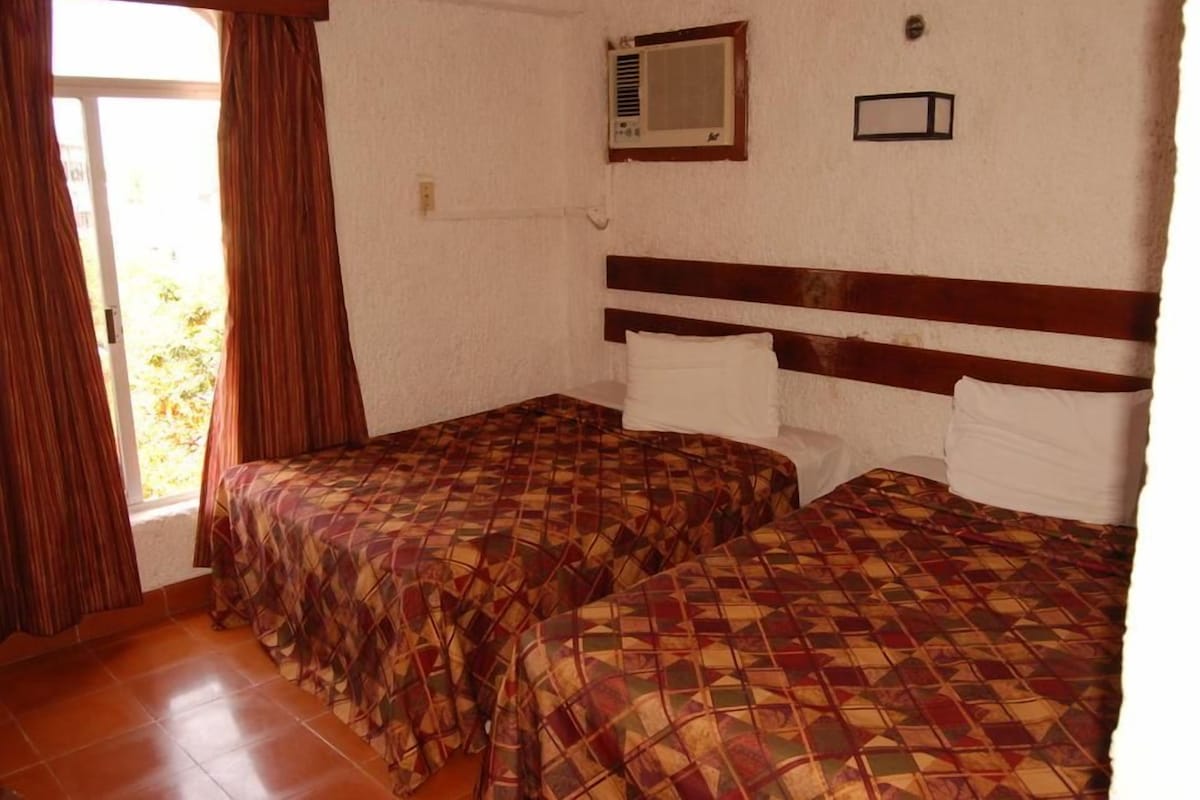 A room with two beds, a window with curtains, an air conditioning unit, and a small wall-mounted light.