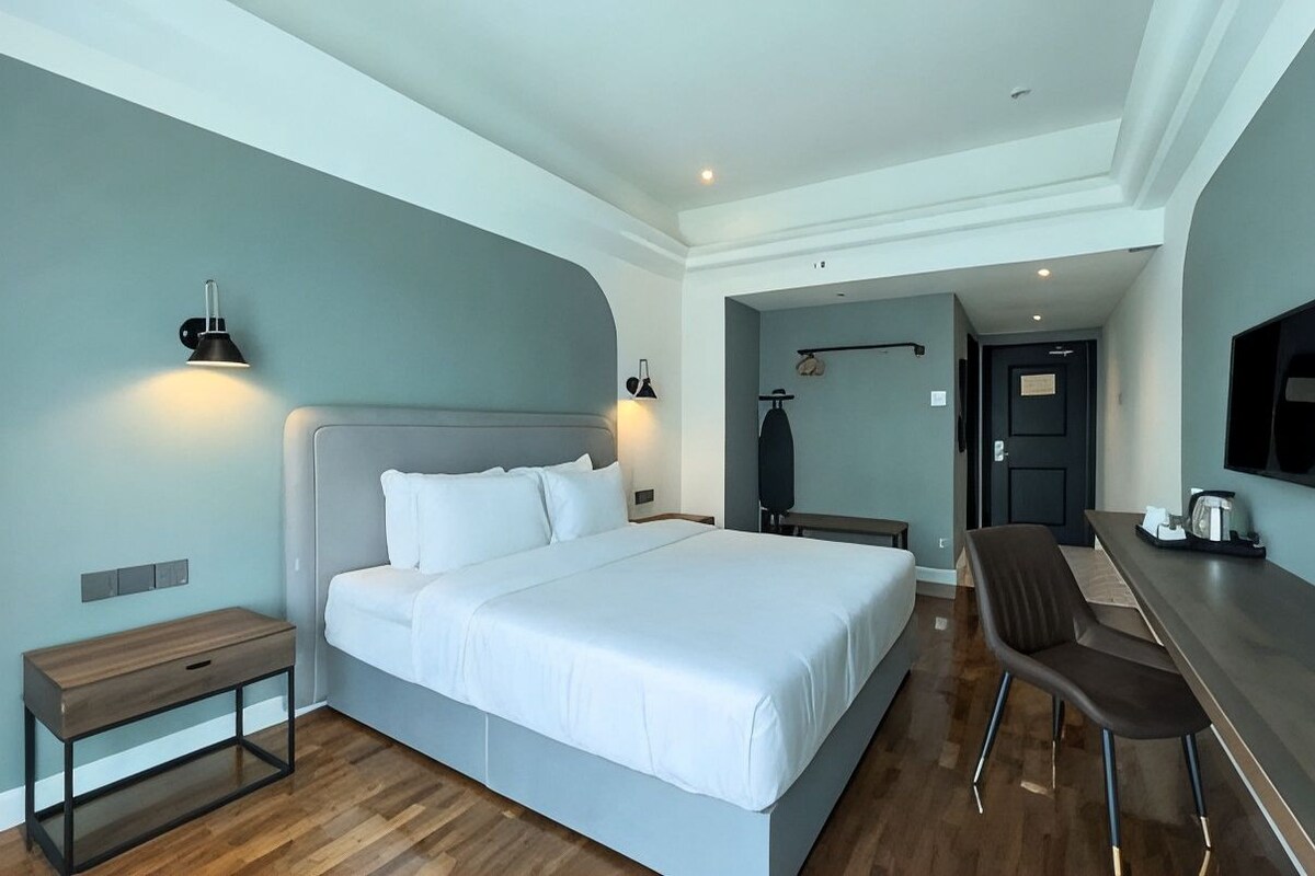 Quarto de hotel moderno com uma cama grande, mesas de cabeceira, escrivaninha e uma cadeira, e uma televisão.