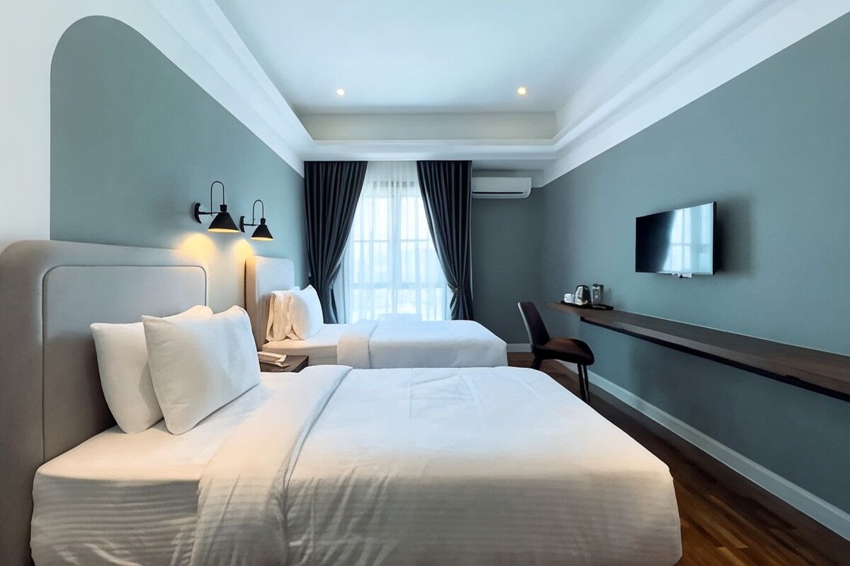 Una habitación de hotel con una cama grande, una mesita de noche, una silla, un televisor montado en la pared y una ventana con cortinas.