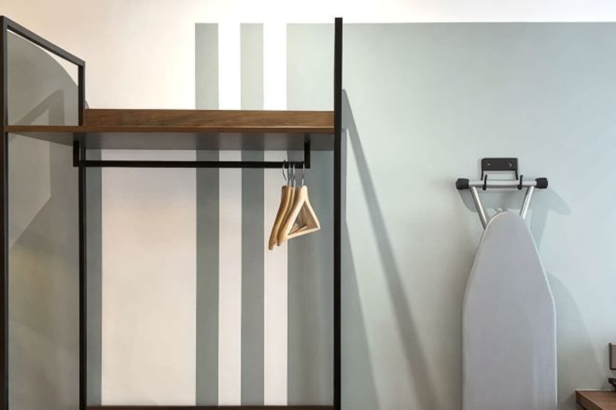 Una habitación con estante de madera, perchero y tabla de planchar blanca frente a una pared con rayas verticales.
