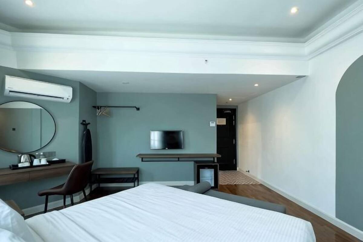 A modern hotel room with a bed, desk, chair, TV, and air conditioning unit.