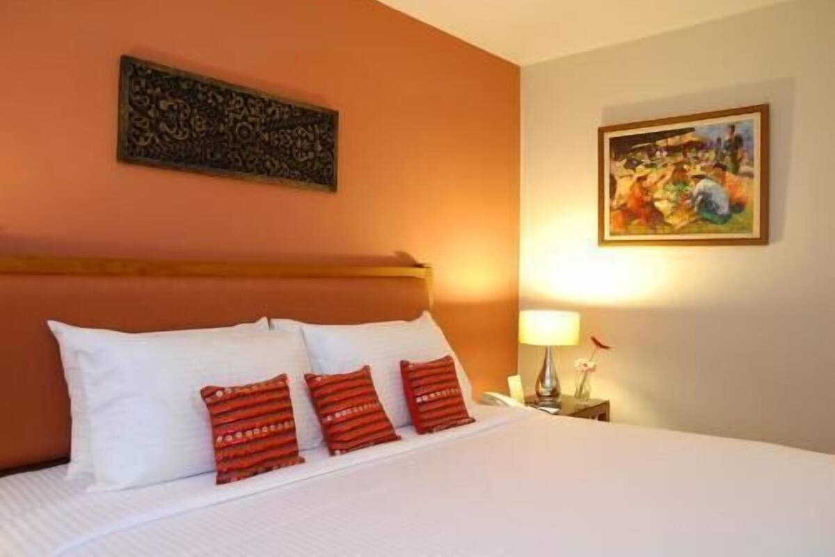 A hotel room with a bed, a headboard, a painting, and a nightstand with a lamp.