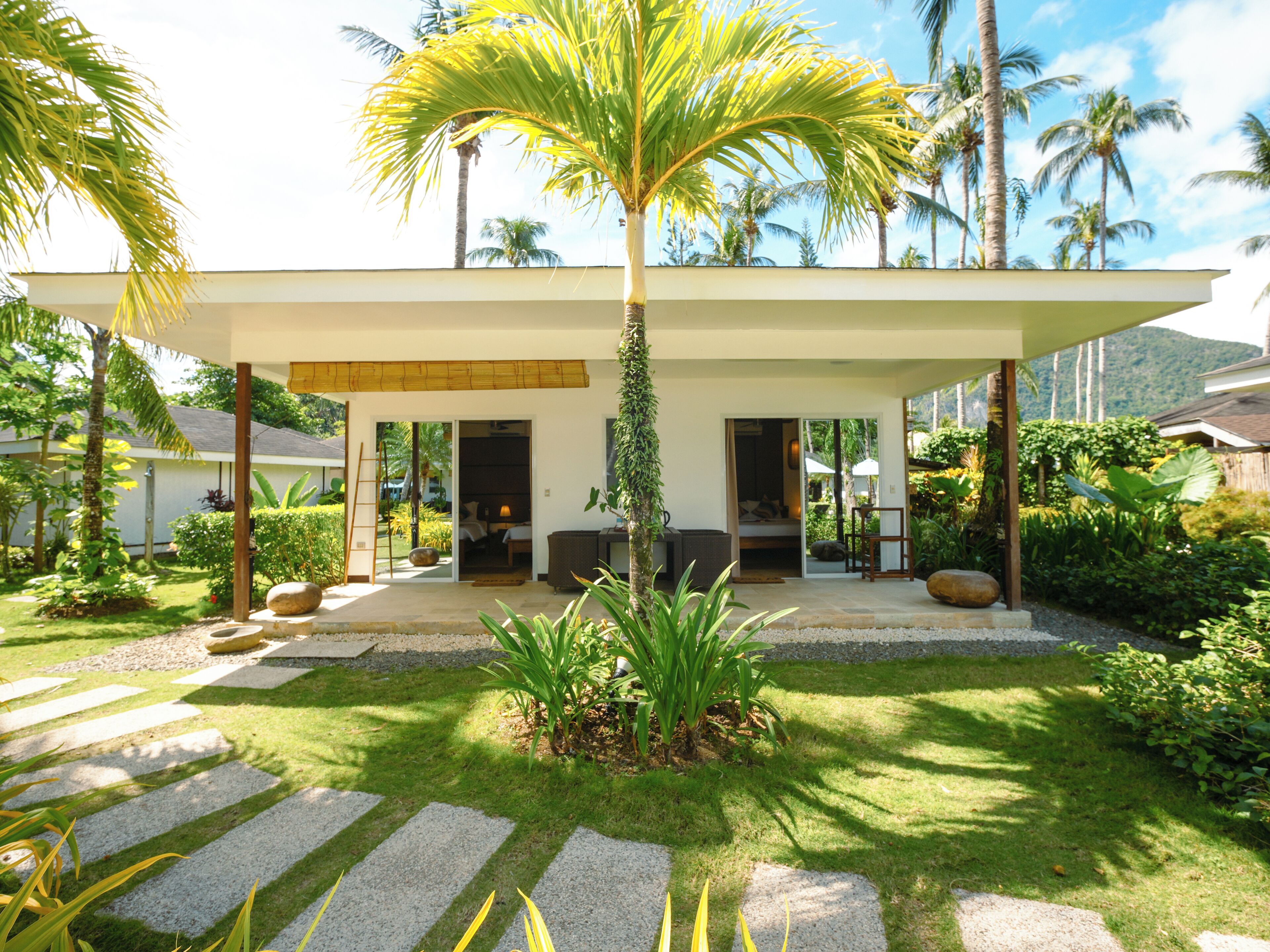 A modern house with a covered patio, a garden with palm trees, and a view of the mountains.