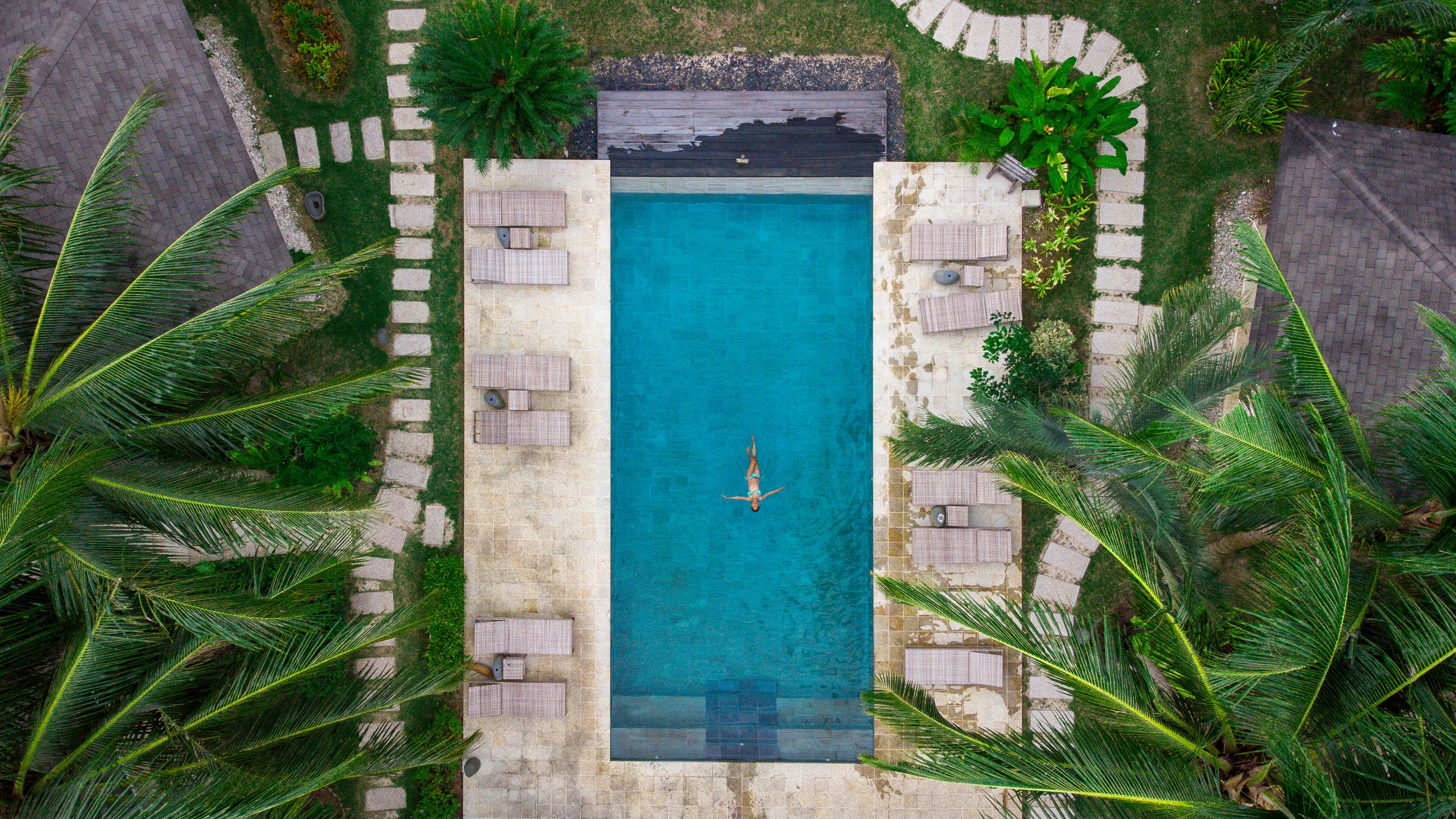 An aerial view of a rectangular swimming pool surrounded by stone steps and palm trees.