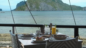 Une table dressée avec des assiettes de nourriture, un verre de jus de fruits et une tasse, donnant sur une étendue d’eau avec, au loin, une île montagneuse.