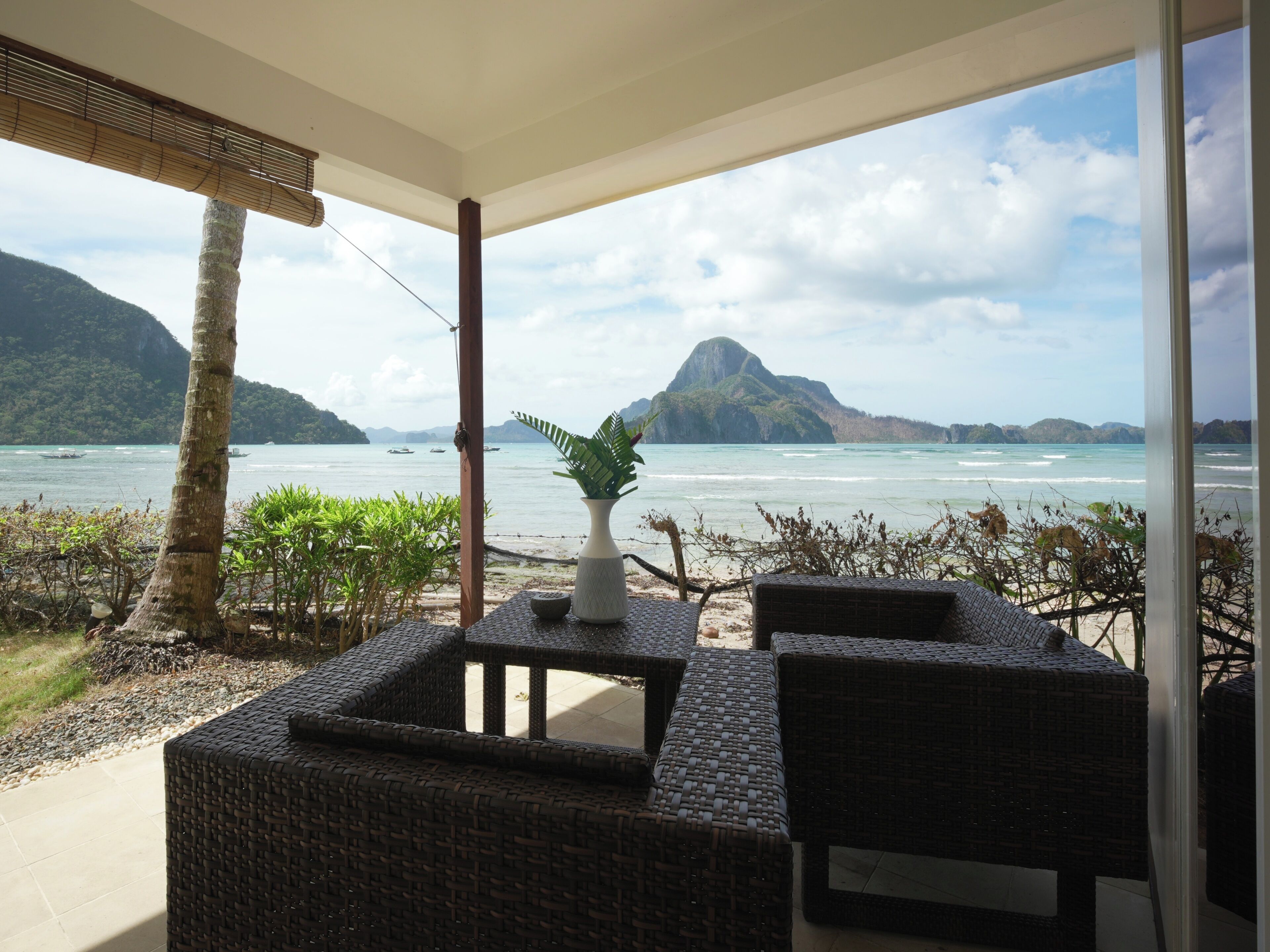 A balcony with wicker furniture, a potted plant, and a view of the sea and mountains.