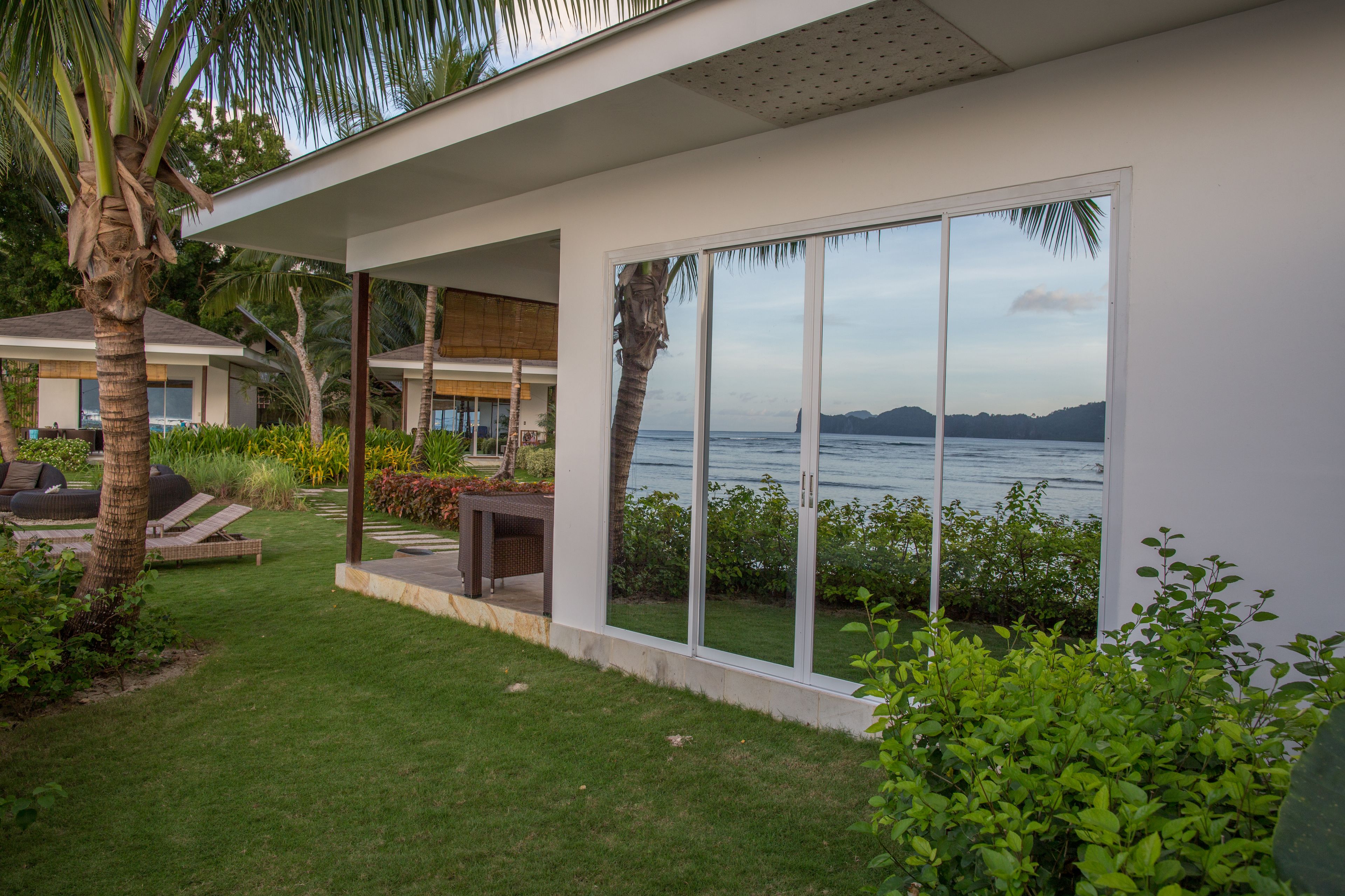 A modern house with a large glass door leading to a patio with outdoor furniture, surrounded by lush greenery and a view of the sea.