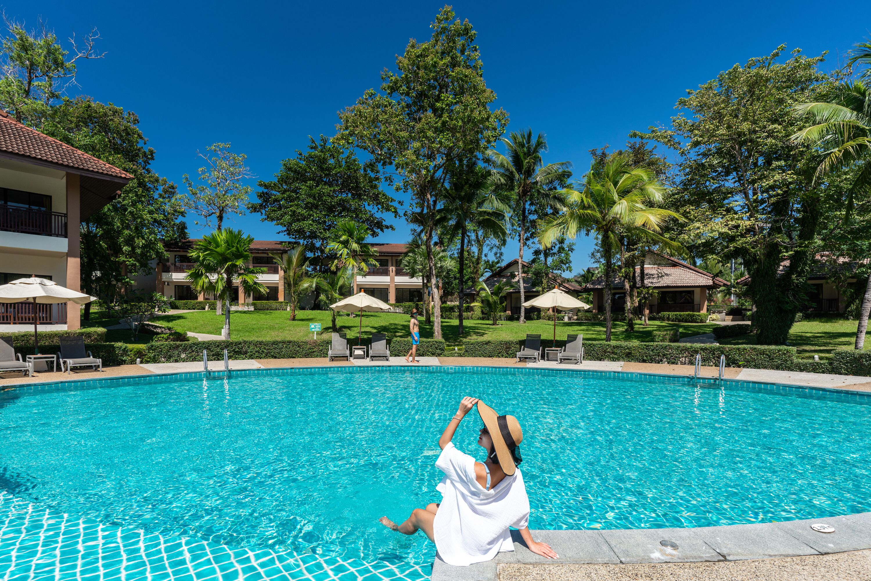 A swimming pool with lounge chairs and umbrellas, surrounded by palm trees and buildings.