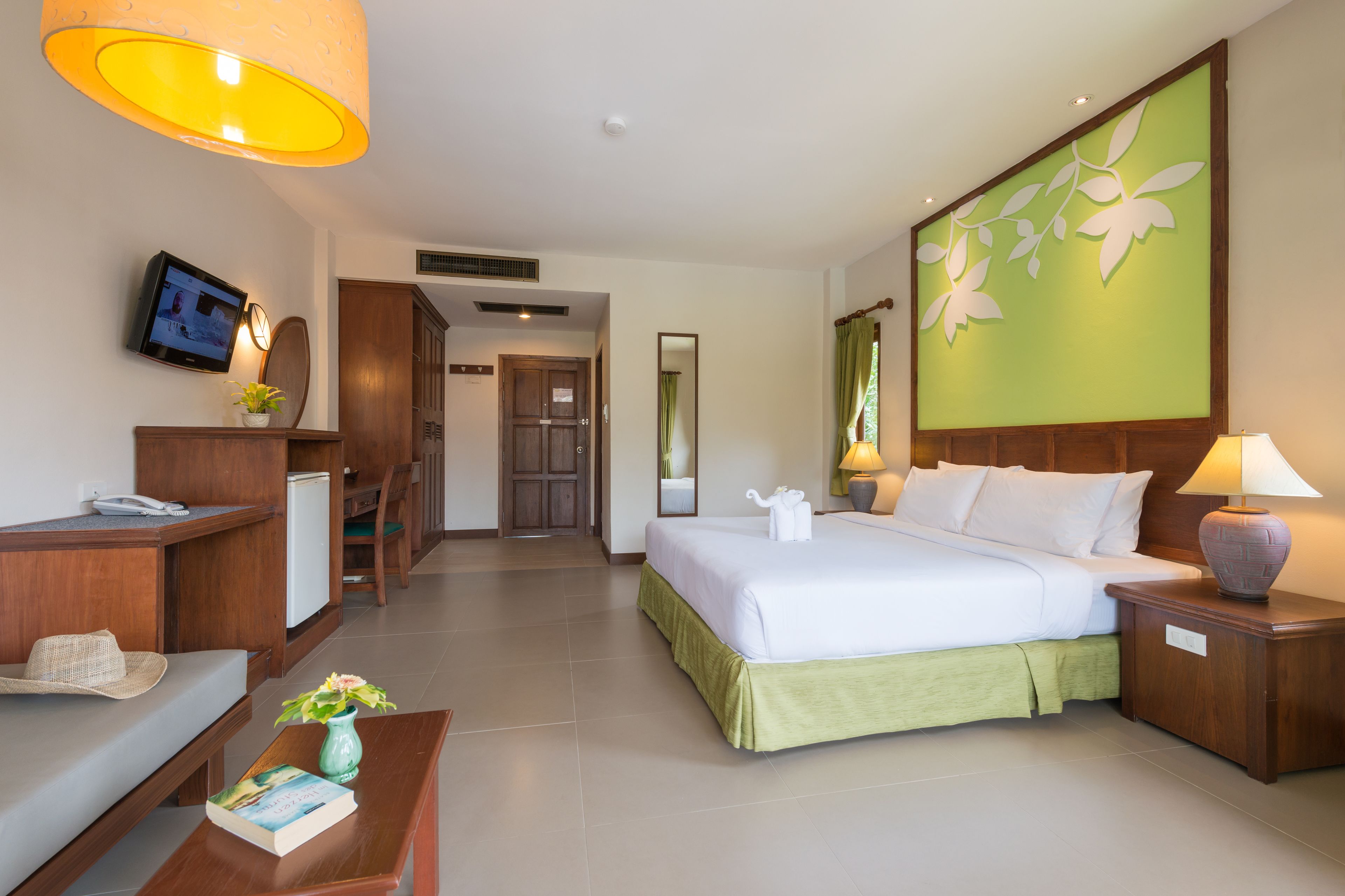 Habitación de hotel con cama, mesita de noche, lámpara, televisor, escritorio, silla y una pequeña mesa con un libro y un jarrón.