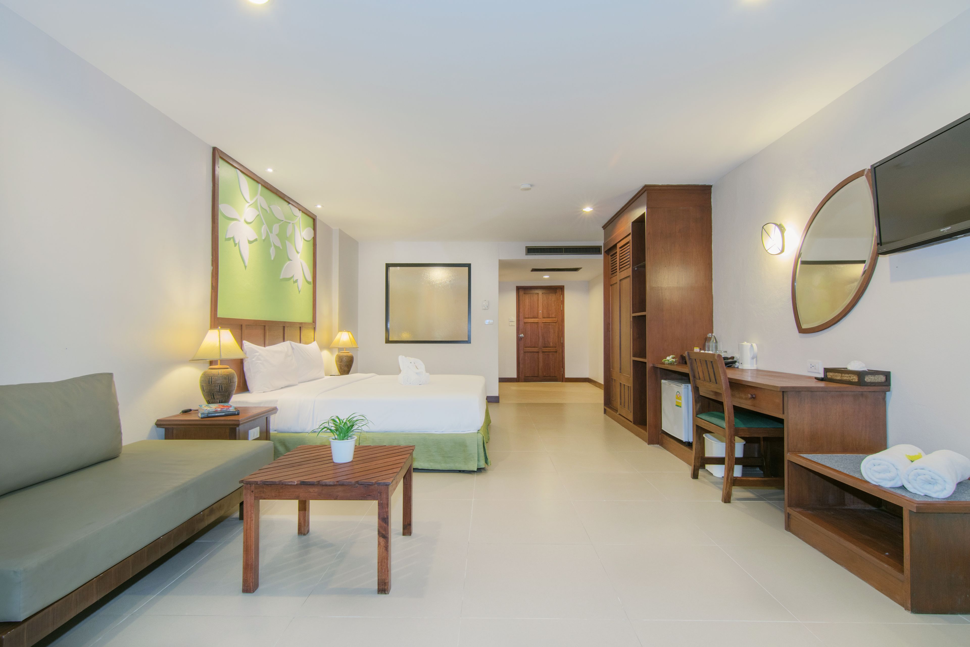 A hotel room with a bed, a sofa, a desk, a chair, a table, a mirror, and a TV.