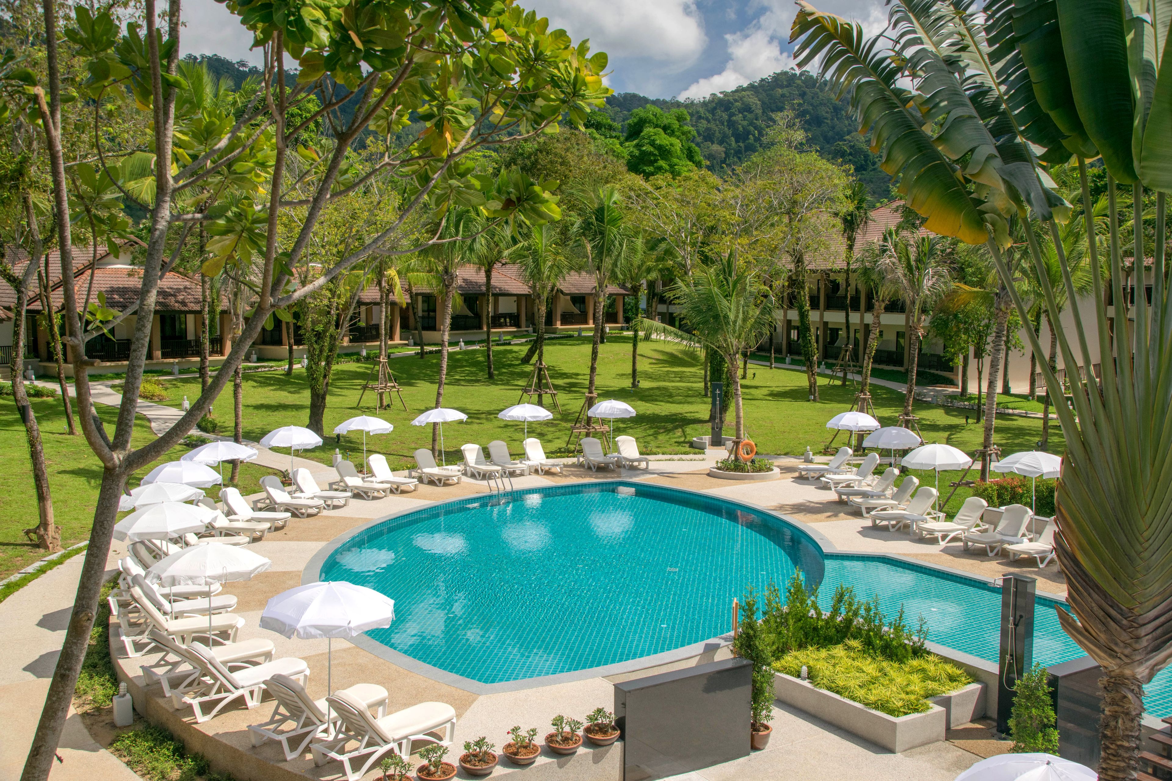 A resort pool area with lounge chairs and umbrellas, surrounded by greenery and a mountain backdrop.
