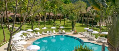 A resort pool area with lounge chairs and umbrellas, surrounded by greenery and a mountain backdrop.