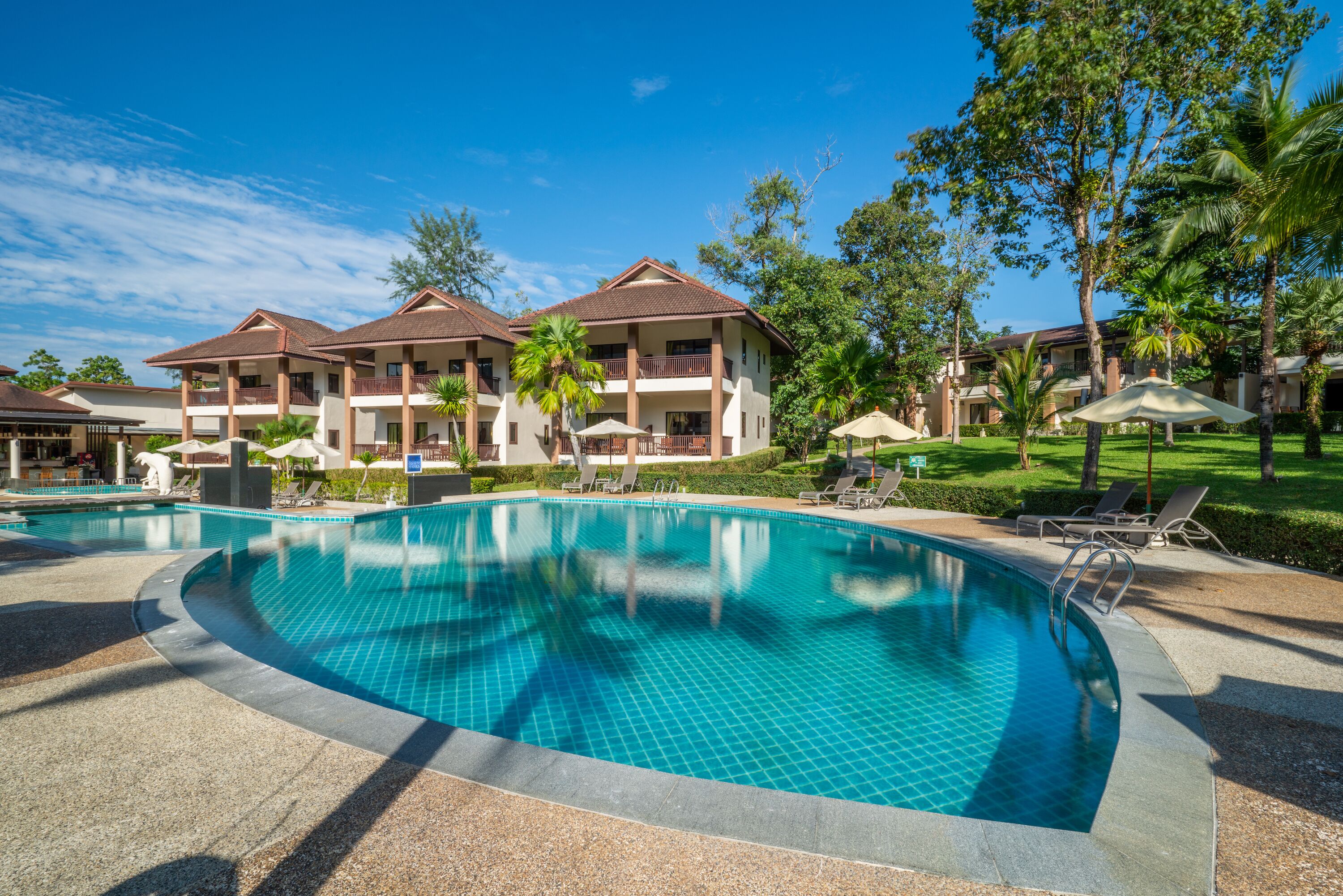 A swimming pool with lounge chairs and umbrellas, surrounded by hotel buildings with balconies and palm trees.