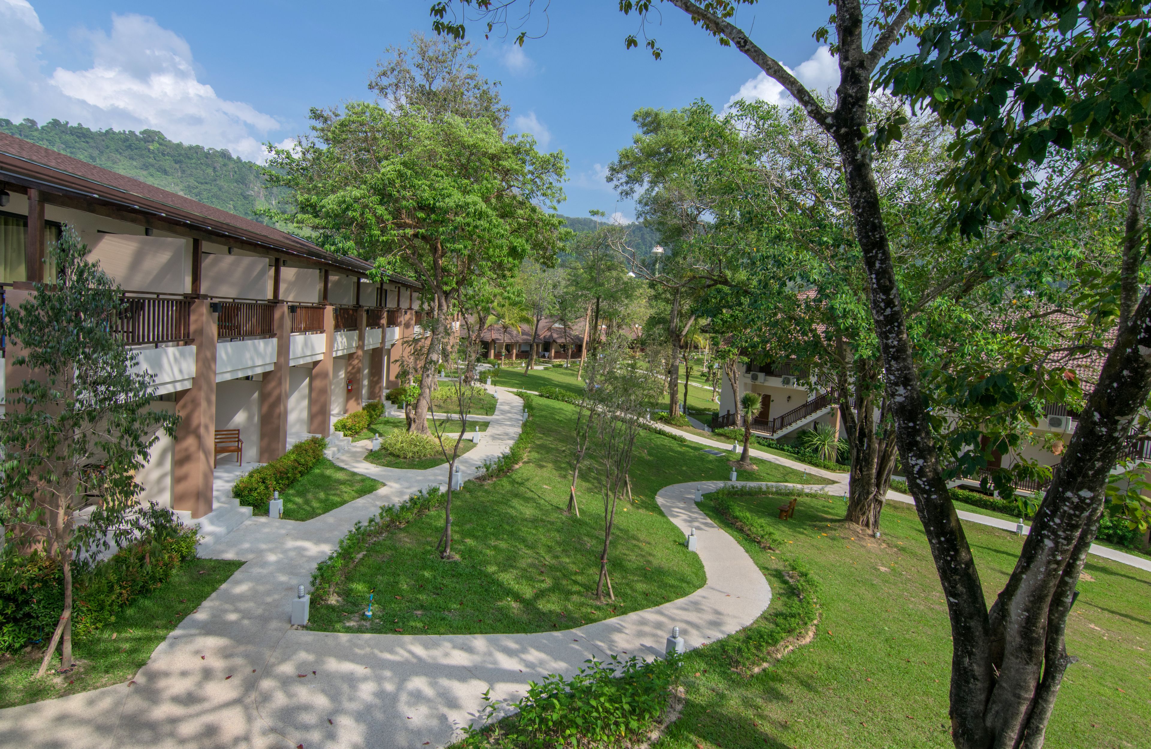 A courtyard with a pathway leading to a building with a balcony. There are trees and a grassy area.