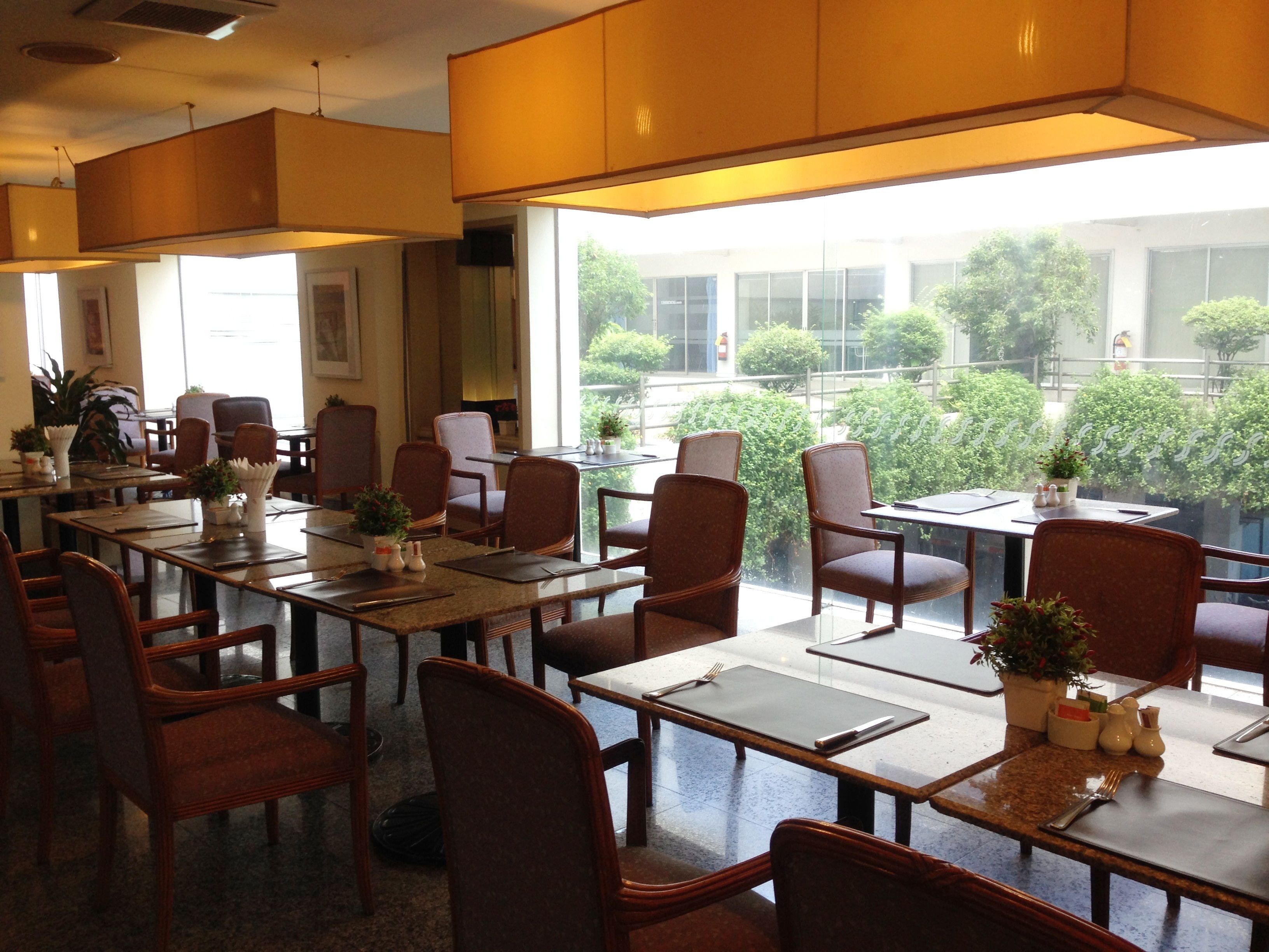 A restaurant with multiple tables and chairs, large windows with a view of greenery outside, and a ceiling with hanging lamps.