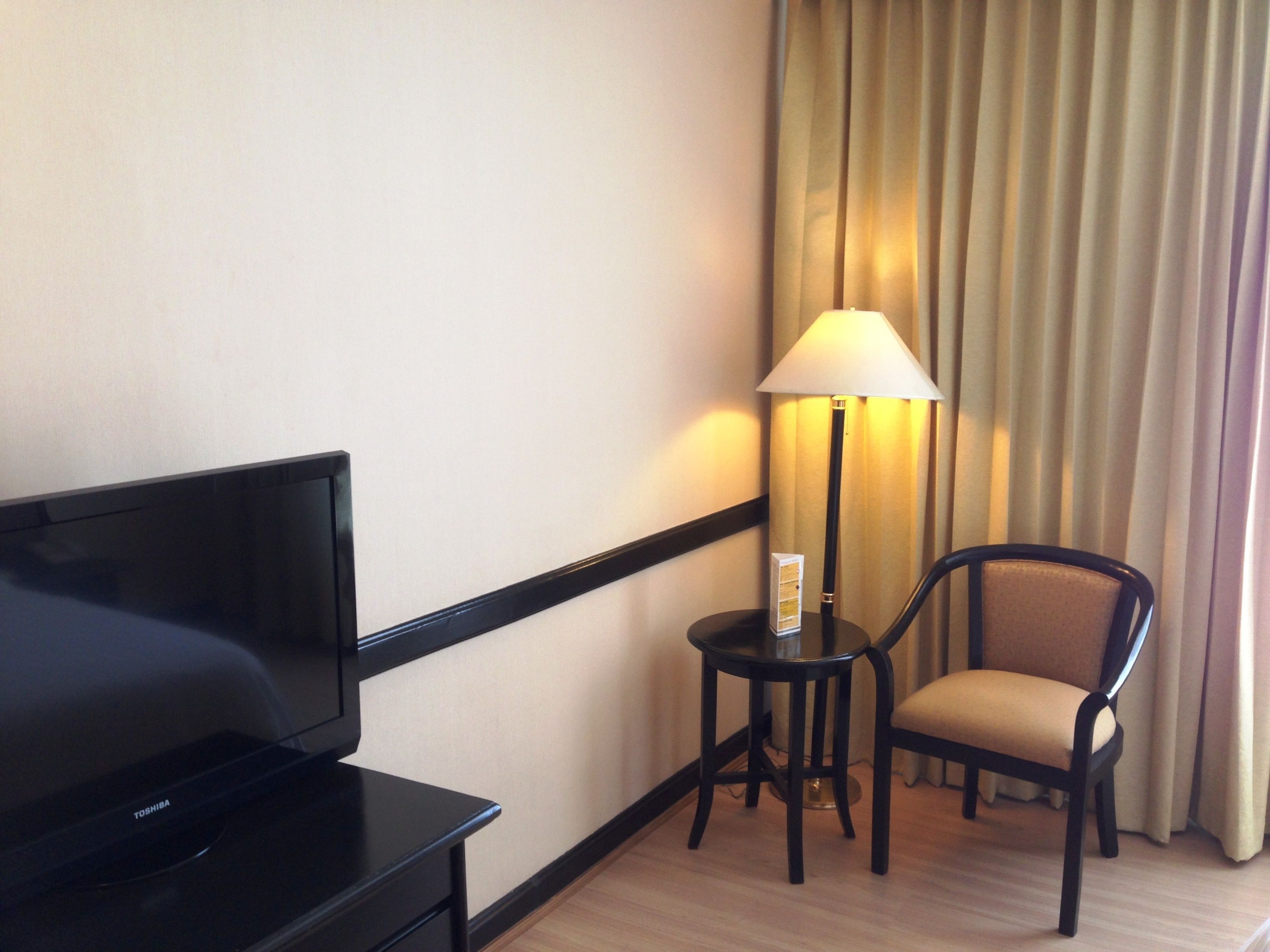 A hotel room with a flat-screen TV, a small table with a lamp, a chair, and a curtain.