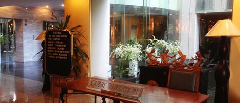 Un vestíbulo de hotel con mostrador de recepción, letrero de atención al huésped, plantas en macetas y un amplio ventanal con vistas a la ciudad.