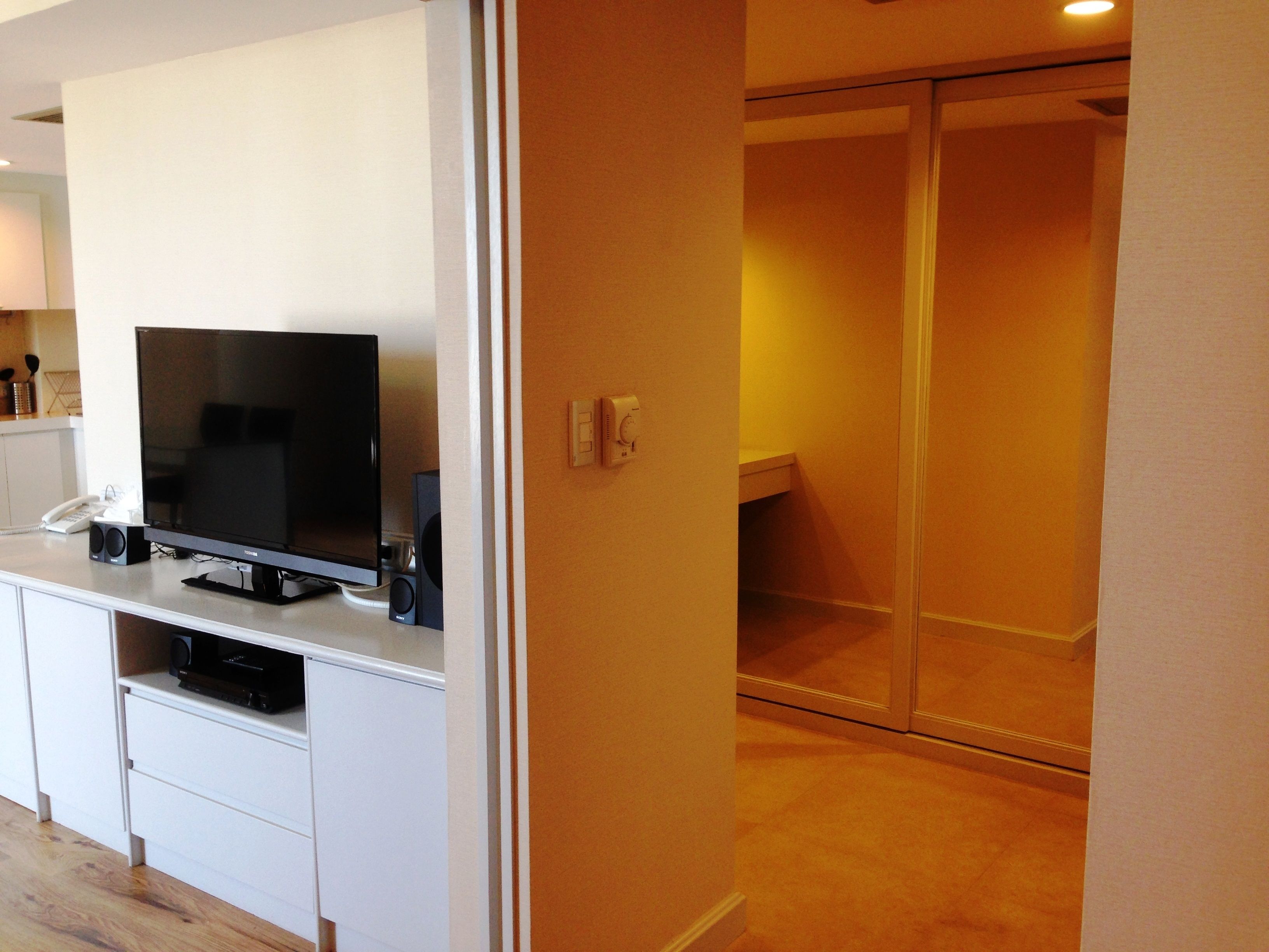A modern hotel room with a flat-screen TV on a white console, a small desk with a telephone, and a sliding door leading to a bathroom.