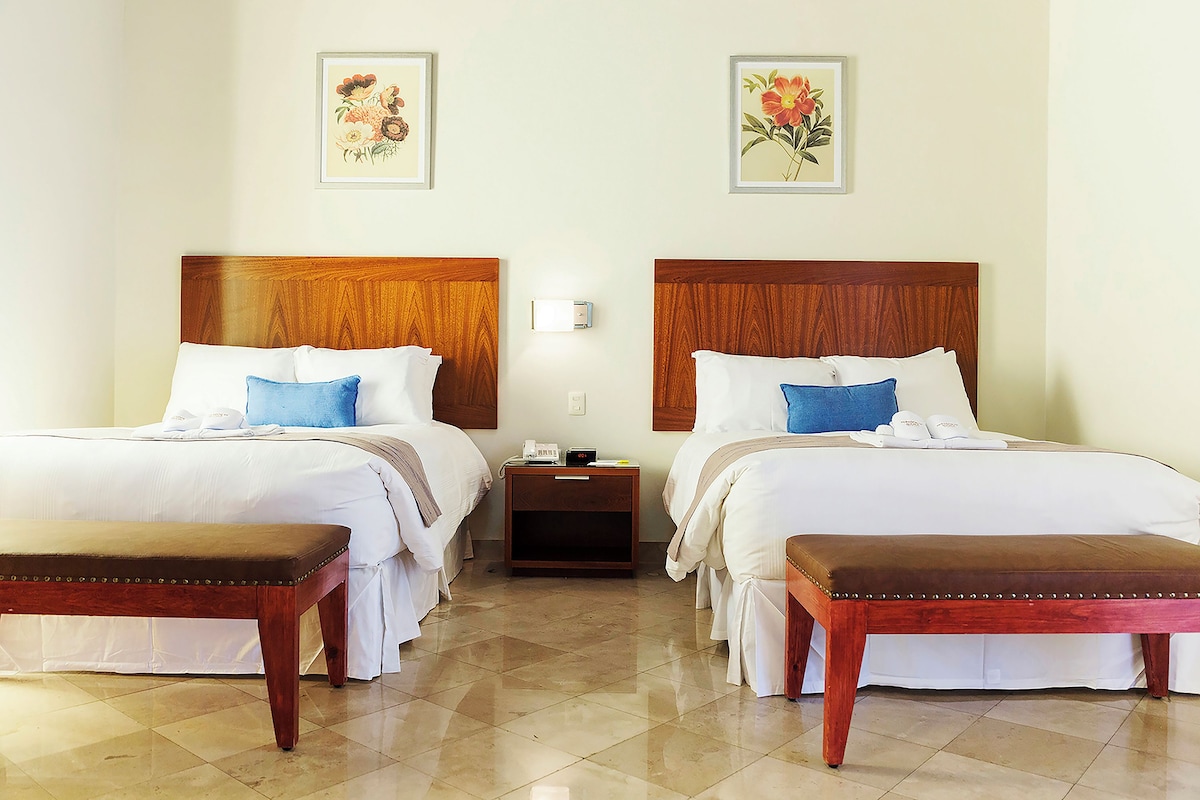 Una habitación de hotel con dos camas, un banco de madera y una mesita de noche con lámpara.