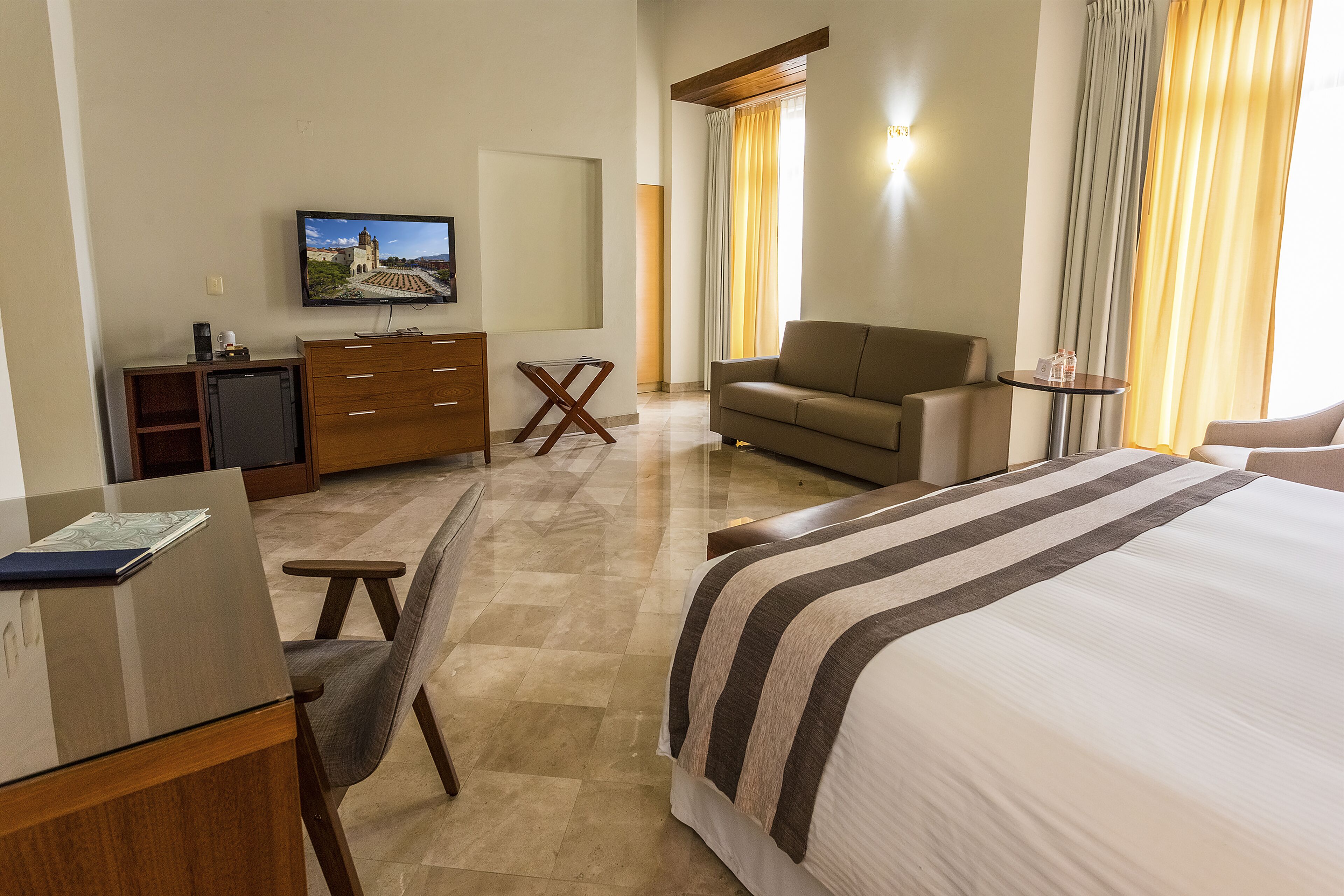 Habitación de hotel con cama, sofá, escritorio con computadora, televisión y una mesita.
