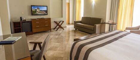 A hotel room with a bed, a sofa, a desk with a computer, a TV, and a small table.