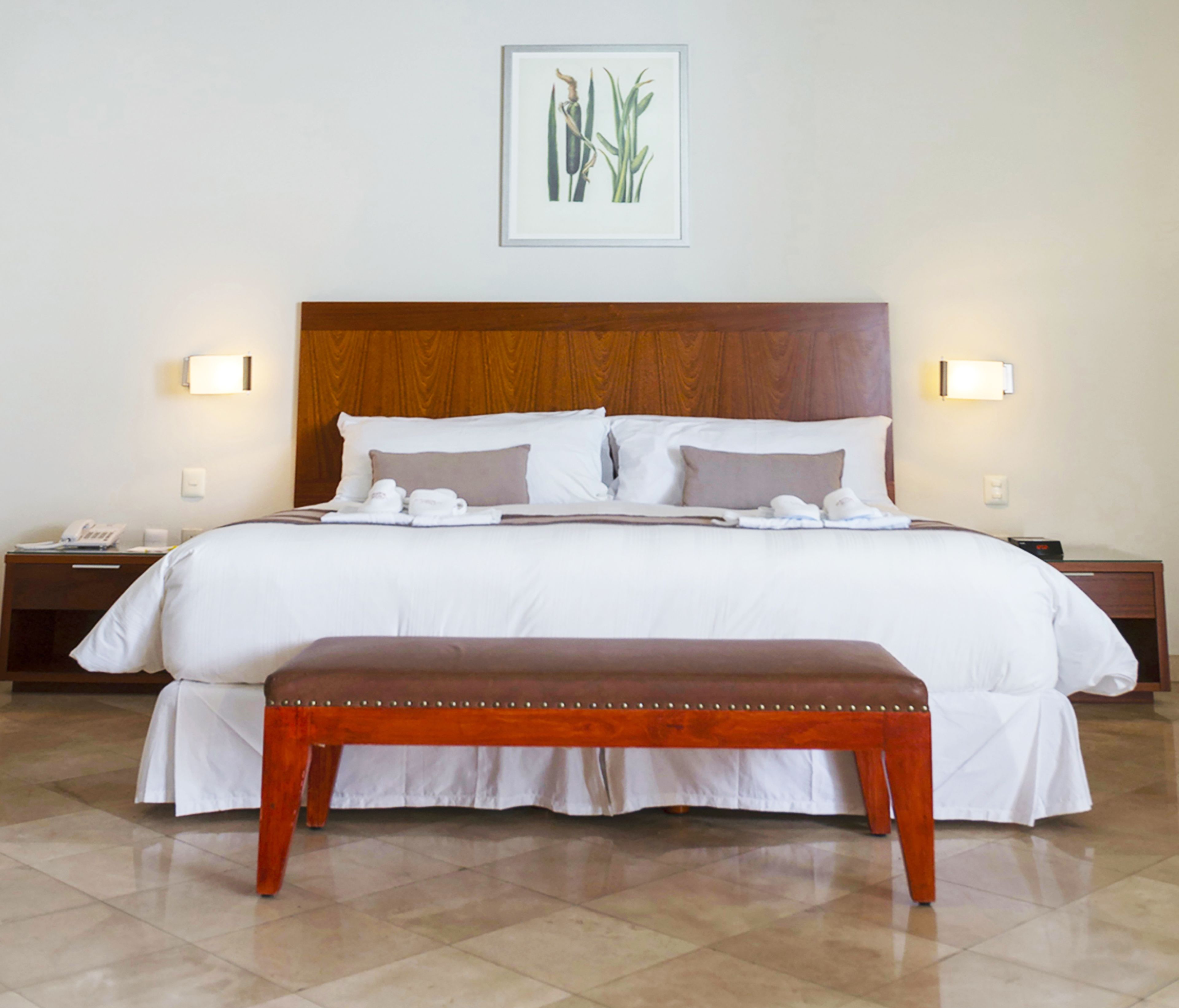 A hotel room with a large bed, two bedside tables, a nightstand, a bench, and a framed botanical illustration on the wall.