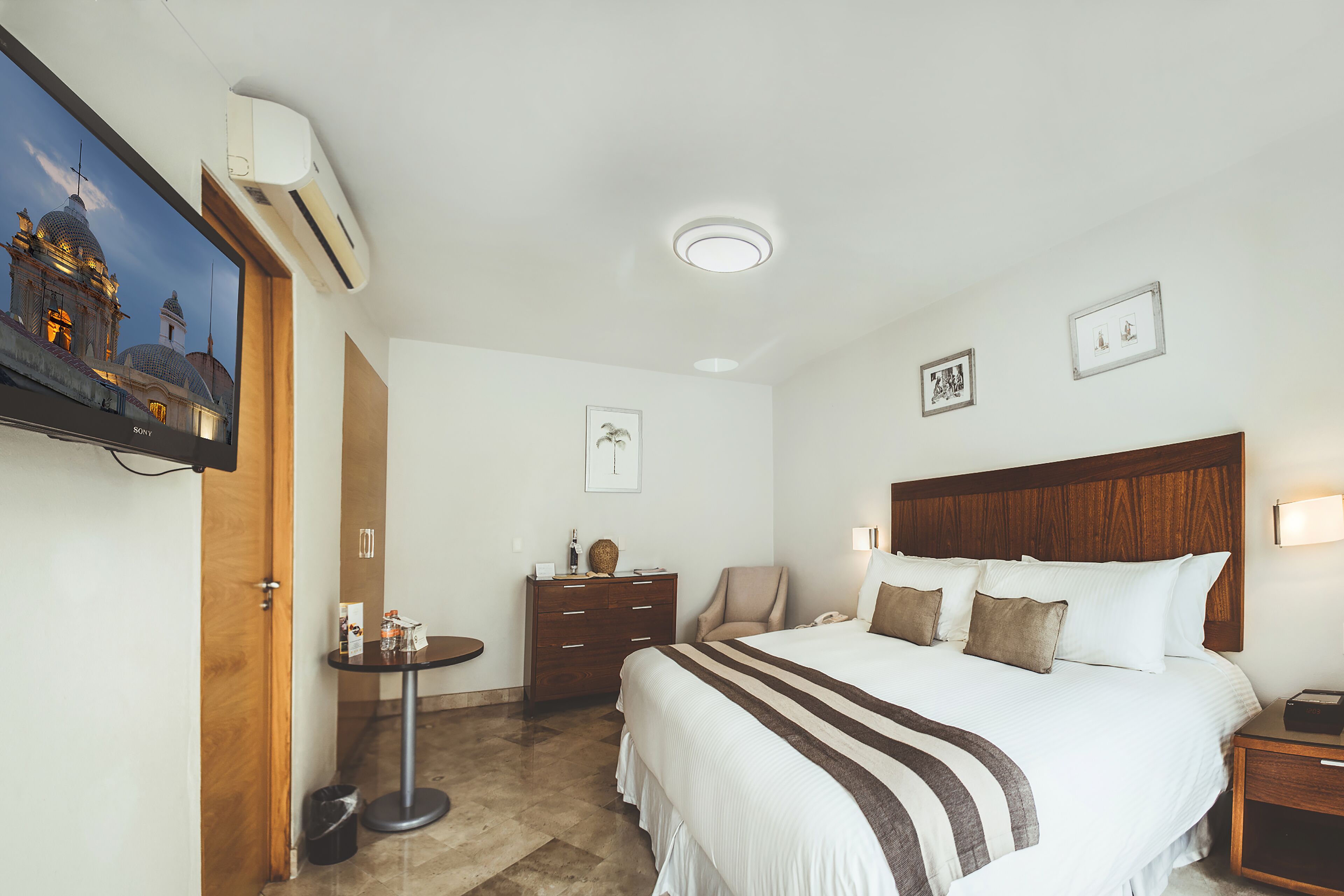 A hotel room with a large bed, a wooden headboard, a bedside table with a lamp, a desk with a chair, and a painting on the wall.