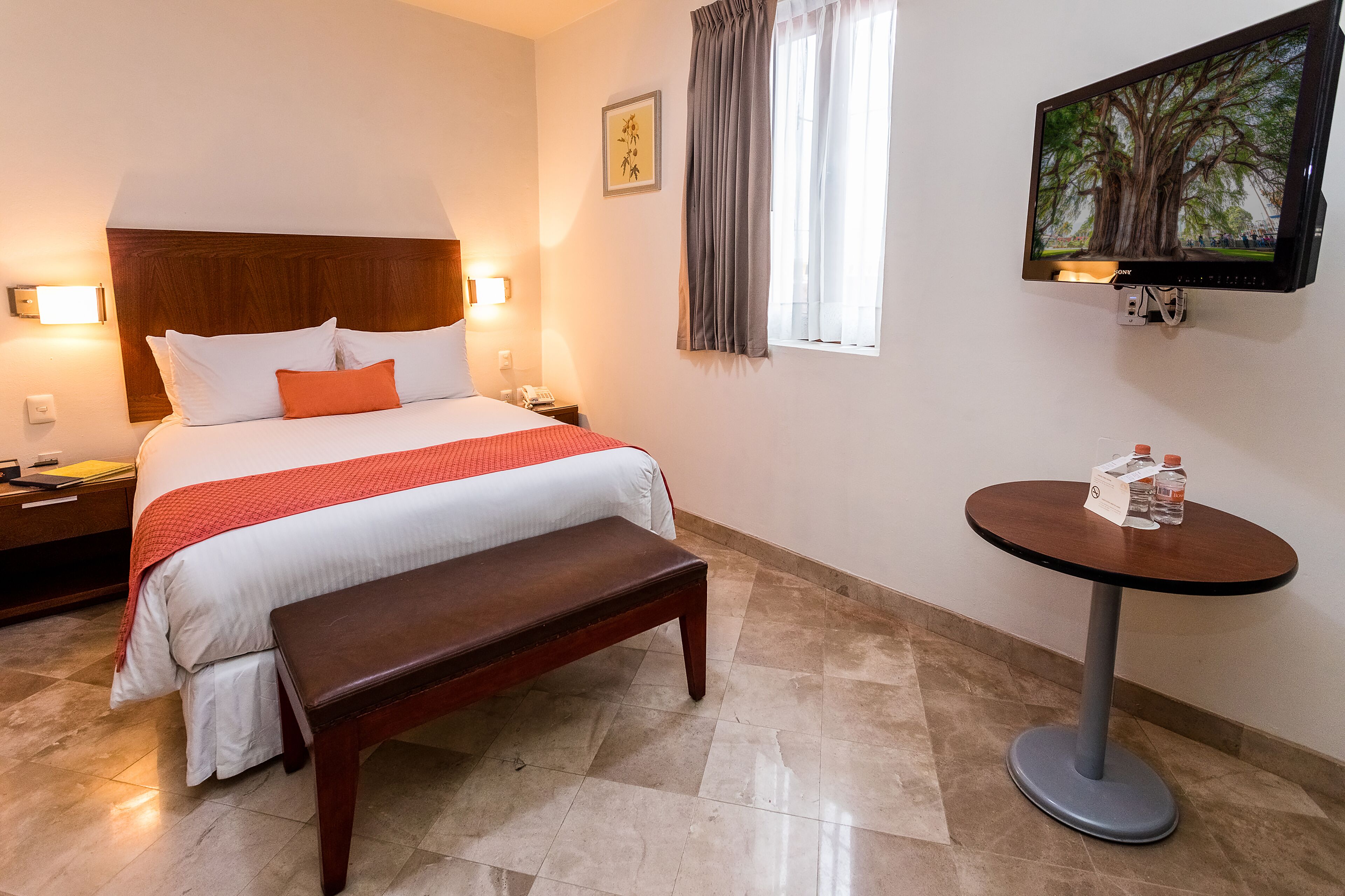 A hotel room with a bed, bench, bedside table, lamp, and a small table with a bottle.
