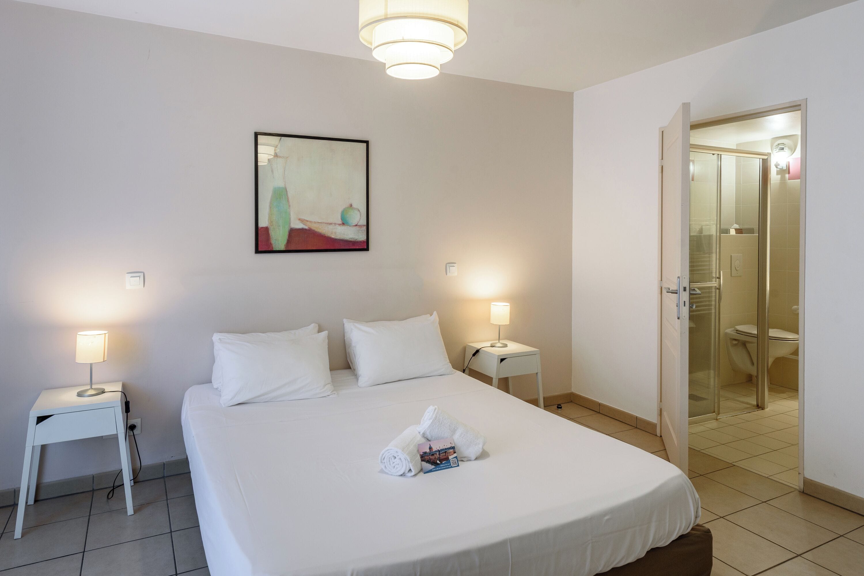 Une chambre d’hôtel avec un lit bien fait, deux lampes de chevet, une petite table avec une lampe, un tableau au mur et une porte en verre pour la douche.