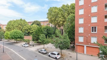 Un edificio de varios pisos de ladrillo con ventanas de marco blanco, un estacionamiento con autos y árboles a lo largo de la acera.