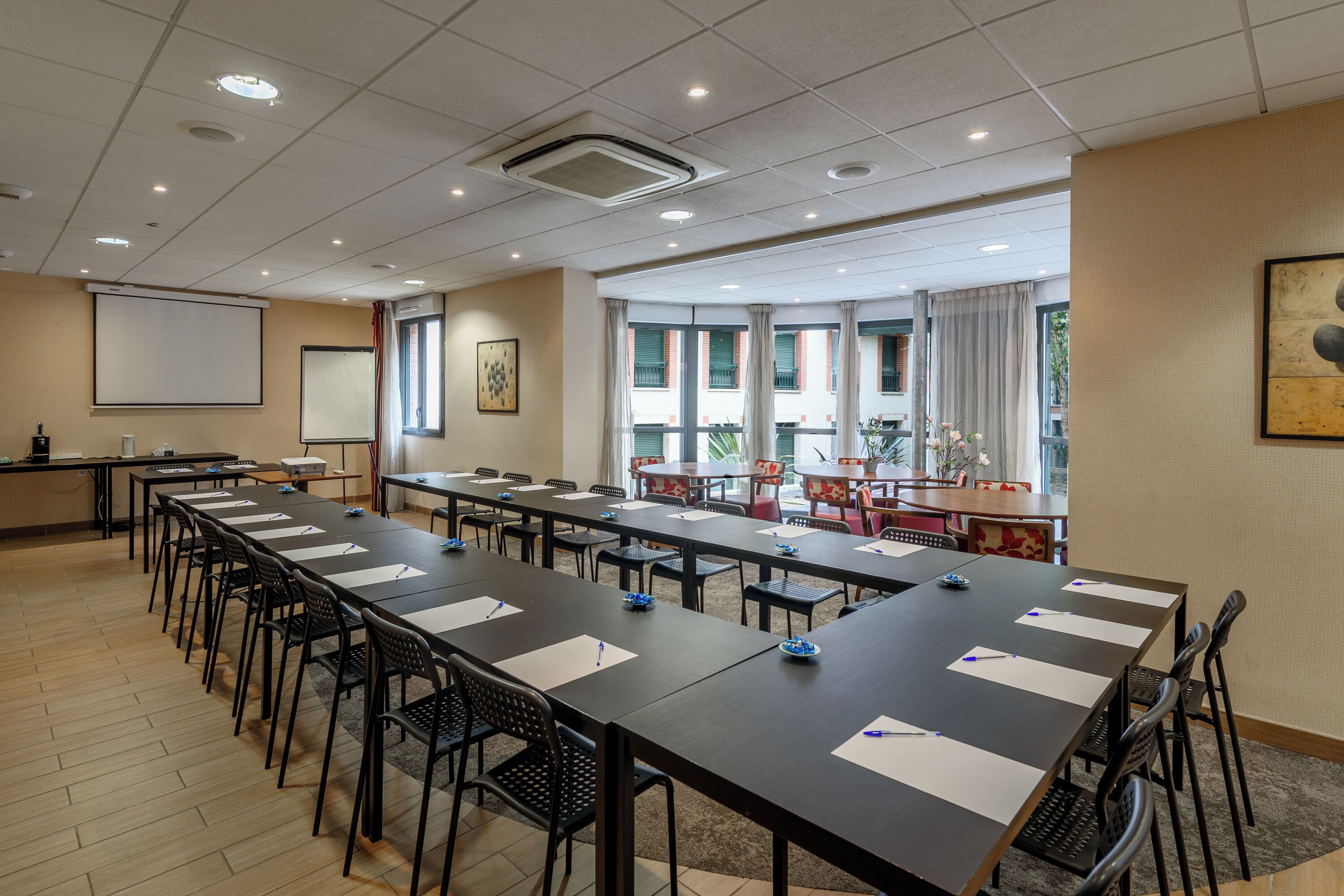 Un salón de conferencias con mesas largas, sillas, una pantalla de proyector y un amplio ventanal con cortinas.