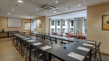 Un salón de conferencias con mesas largas, sillas, una pantalla de proyector y un amplio ventanal con cortinas.