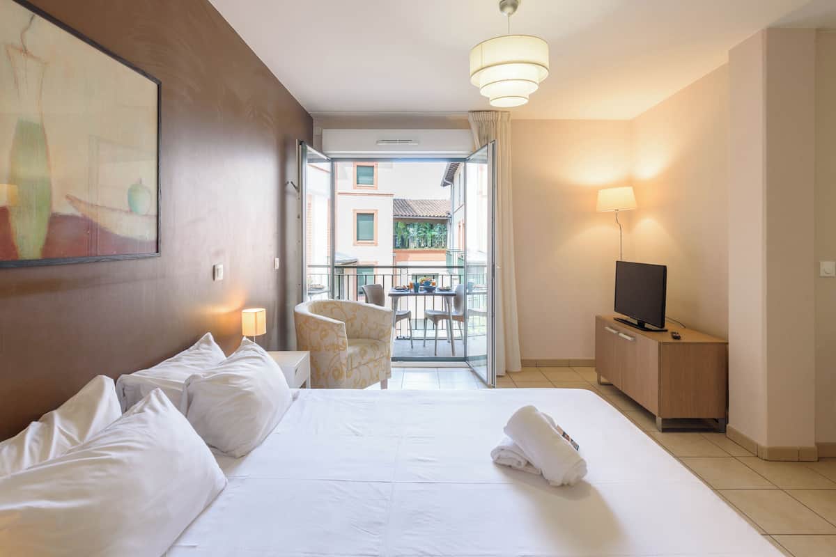 Habitación de hotel con cama, televisión sobre un mueble de madera, una silla y una mesa con vistas al exterior.