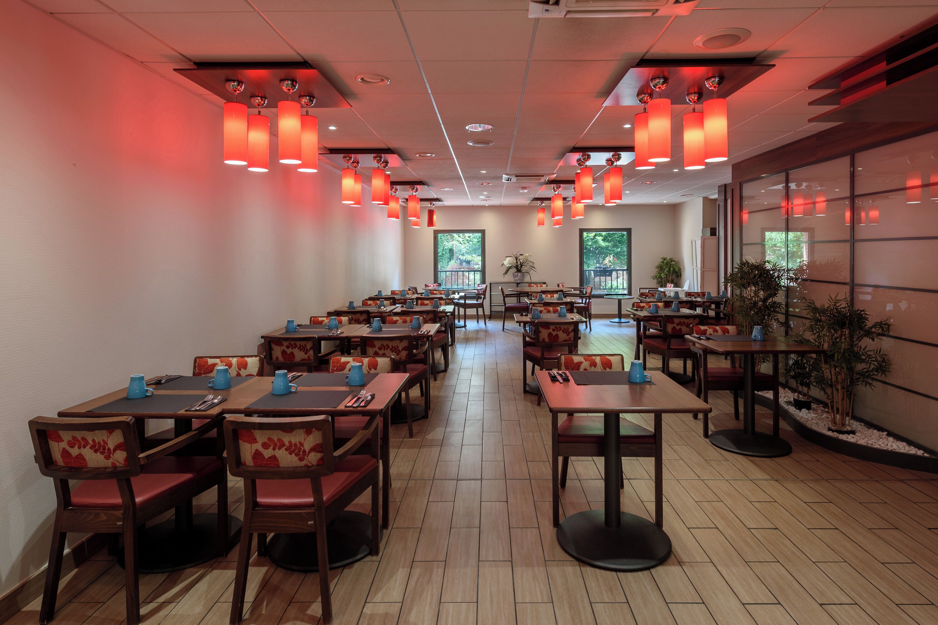 Un restaurante con varias mesas y sillas, luces colgantes rojas y amplios ventanales.
