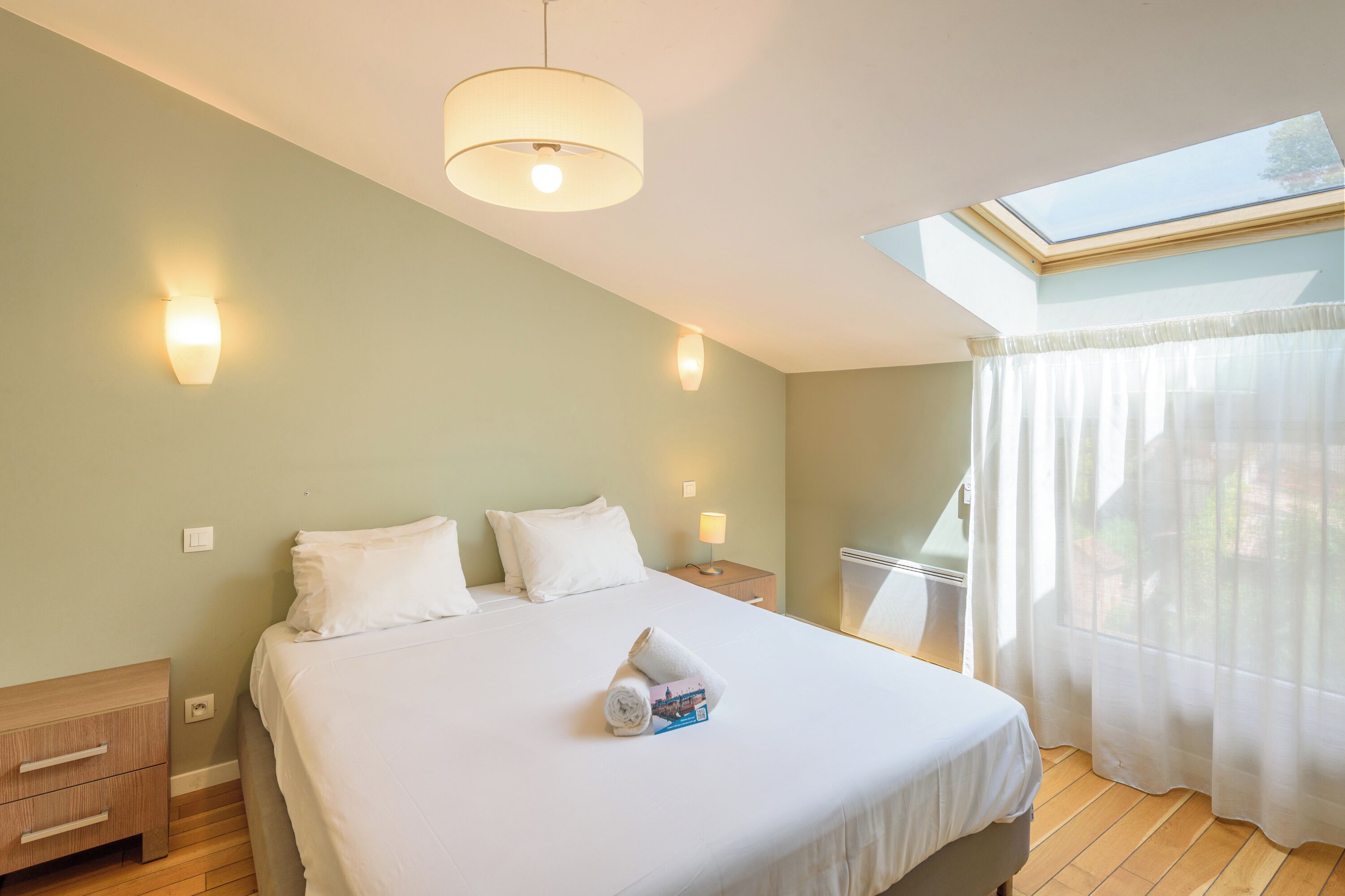 Un dormitorio con tragaluz, una cama con ropa de cama blanca, una mesita de noche con lámpara y una mesita auxiliar de madera.